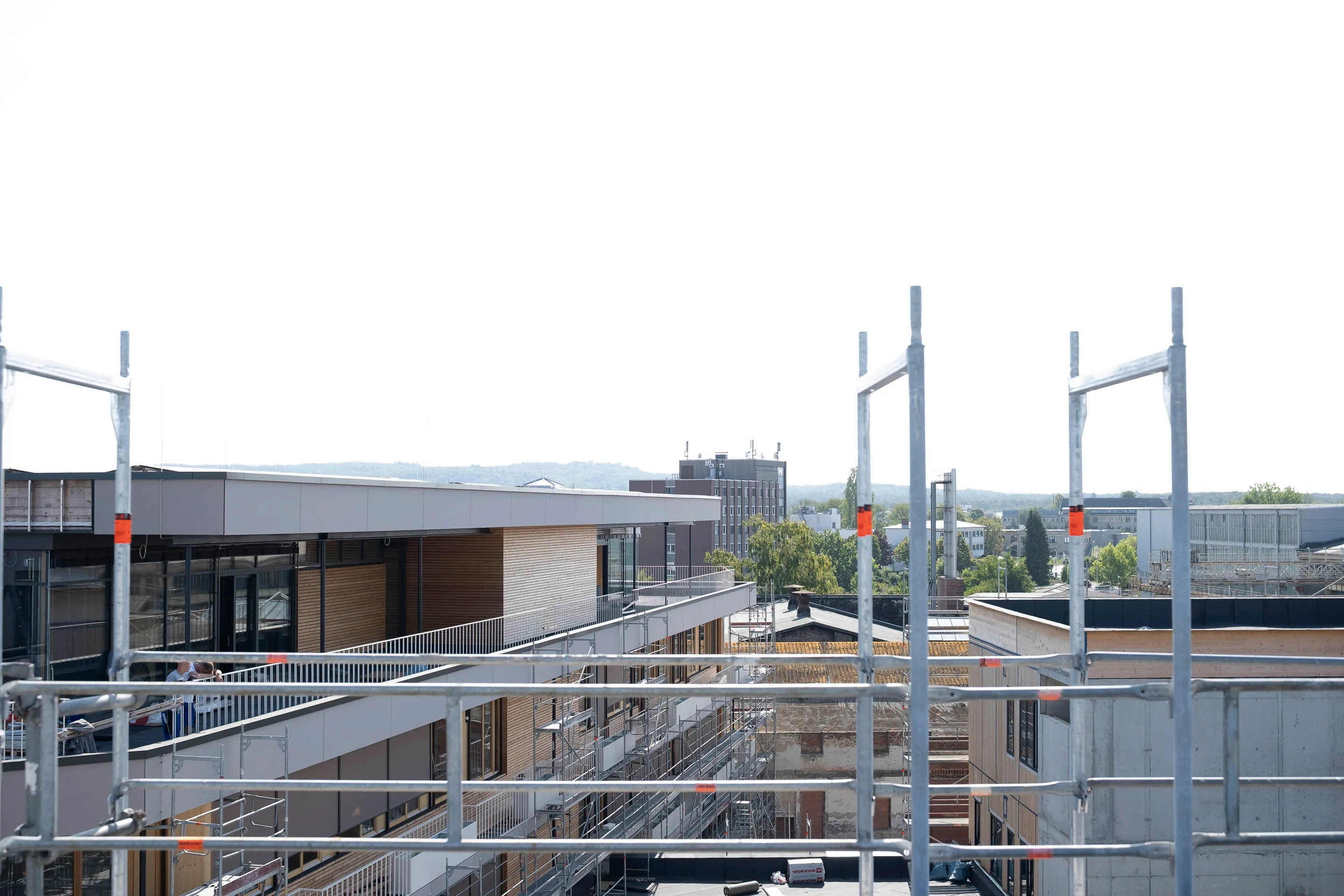 View of modern buildings under construction with metal scaffolding and distant trees under a bright sky.