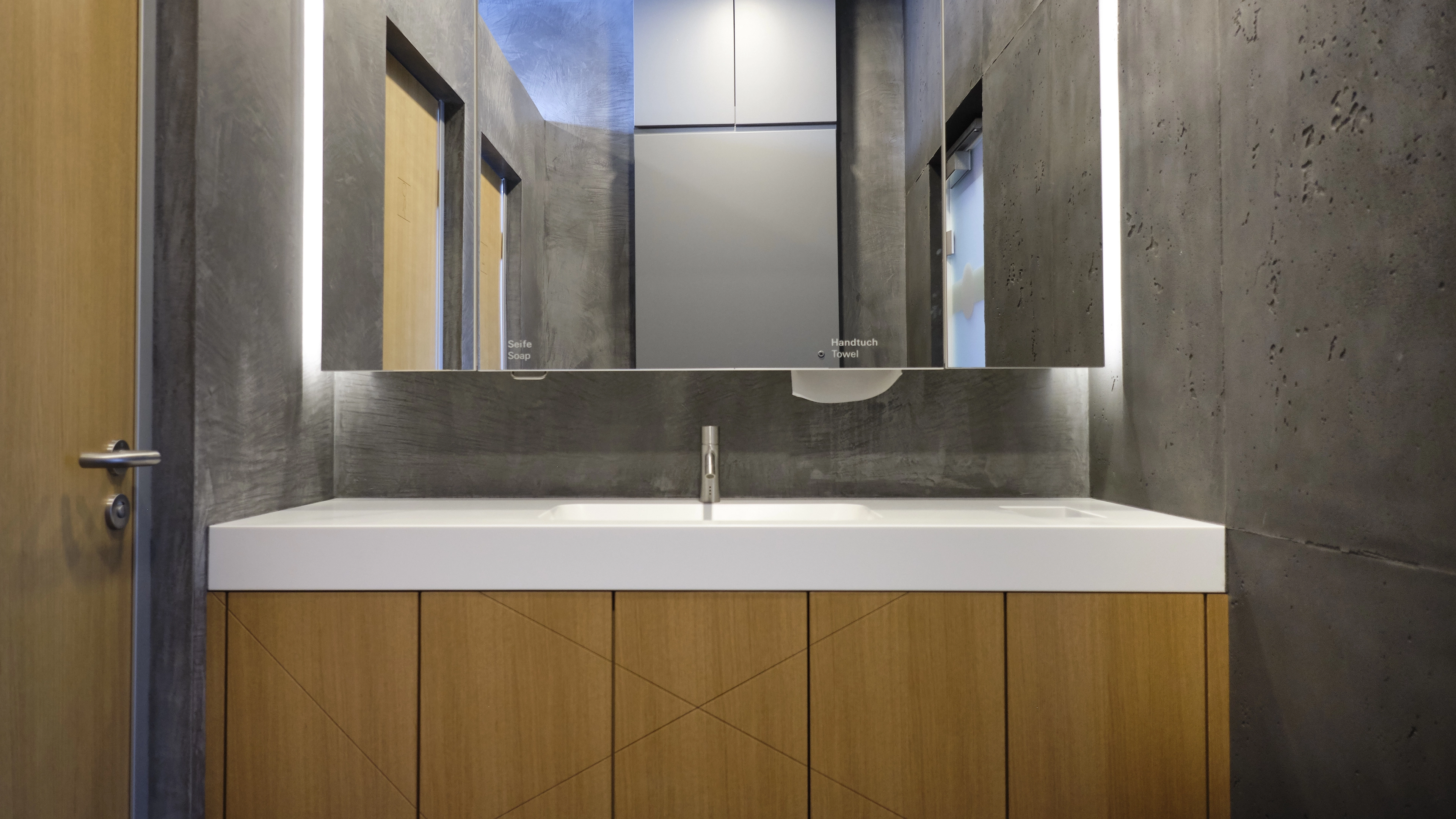 Modern bathroom sink with wooden cabinet, concrete walls, and a large mirror reflecting doors and ceiling.