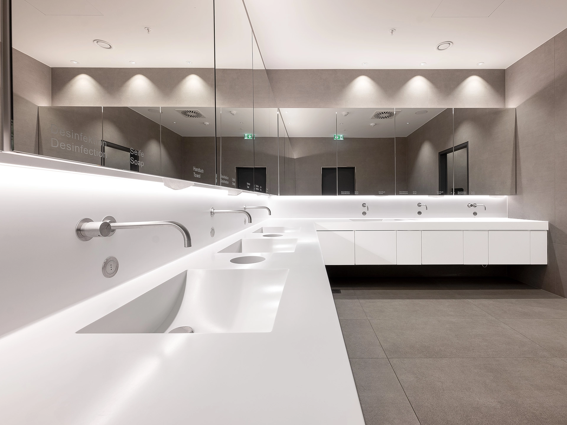 Modern public restroom with white counters, integrated sinks, wall-mounted faucets, large mirrors, and gray tiled walls and floor.