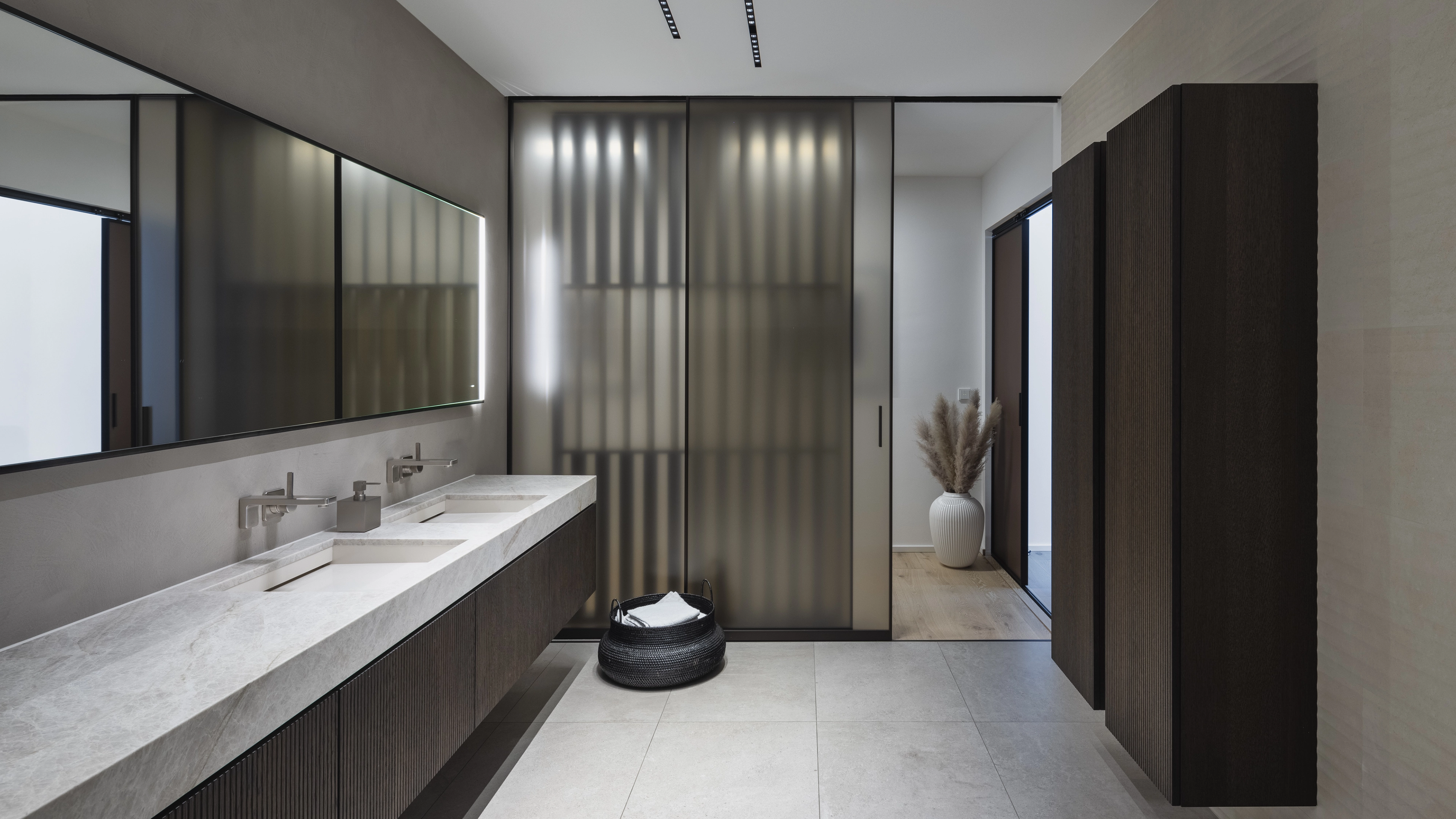 Modern bathroom with double sink vanity, large illuminated mirror, frosted glass sliding doors, and dark wood storage cabinets.