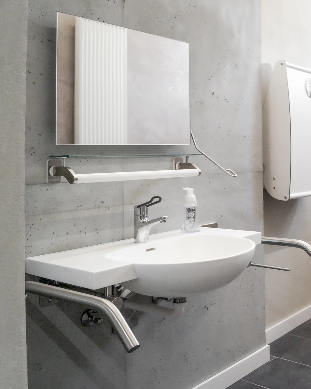 Modern wall-mounted white sink with chrome faucet, mirror, soap dispenser, and support bars in a bathroom with gray tiled walls.