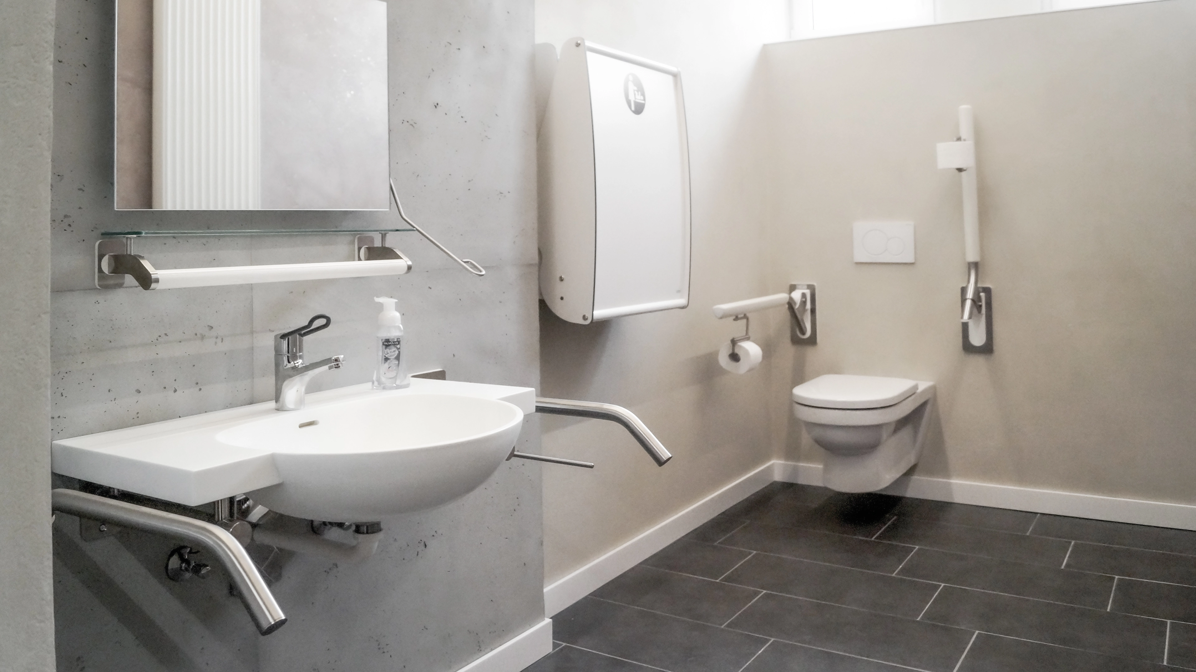 Accessible bathroom with wall-mounted sink and toilet, grab bars, soap dispenser, and changing table.