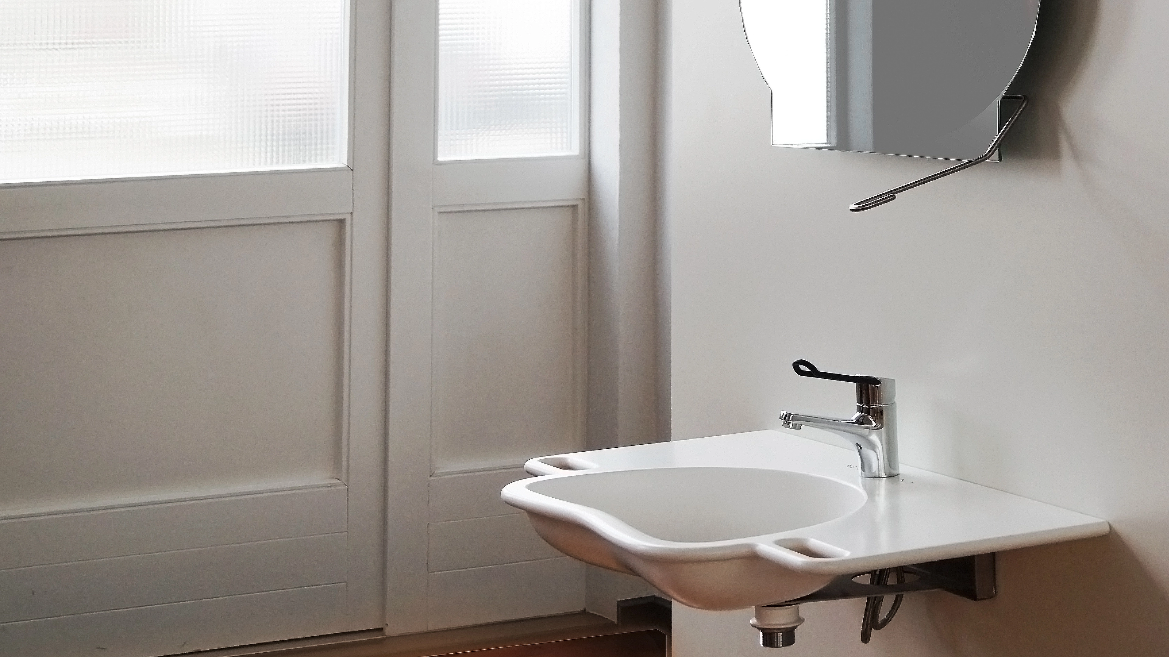 Accessible wall-mounted white bathroom sink with lever faucet and a round mirror above it.