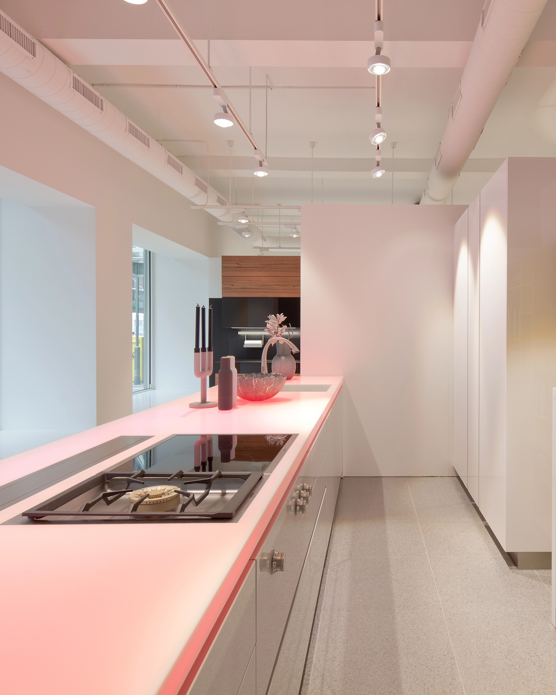 Modern kitchen with a long white island counter featuring an integrated stovetop, sink, and decorative items including candlesticks and a glass bowl under soft pink lighting.