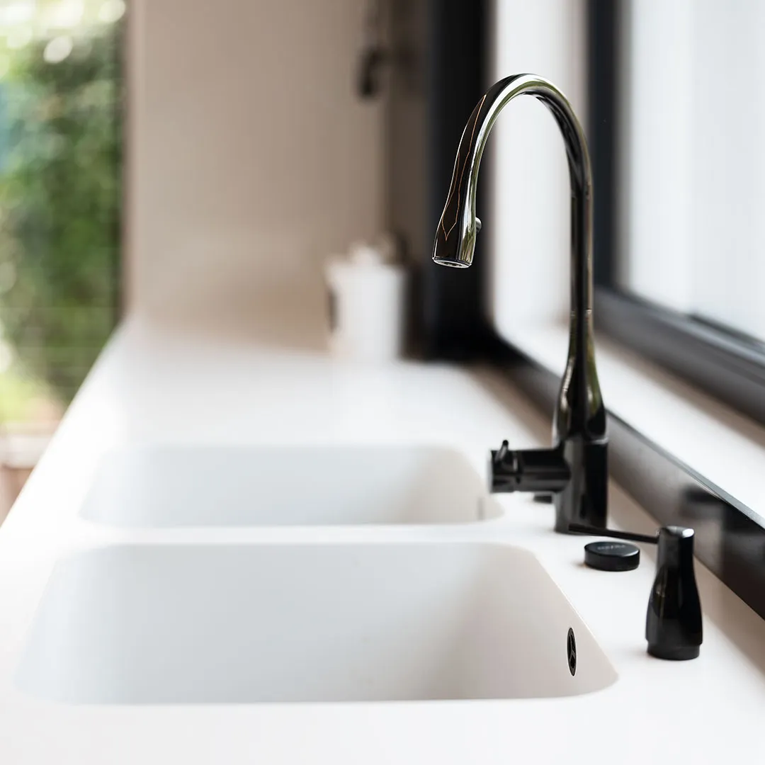 Modern kitchen sink with dual white basins and a sleek black curved faucet next to a window.