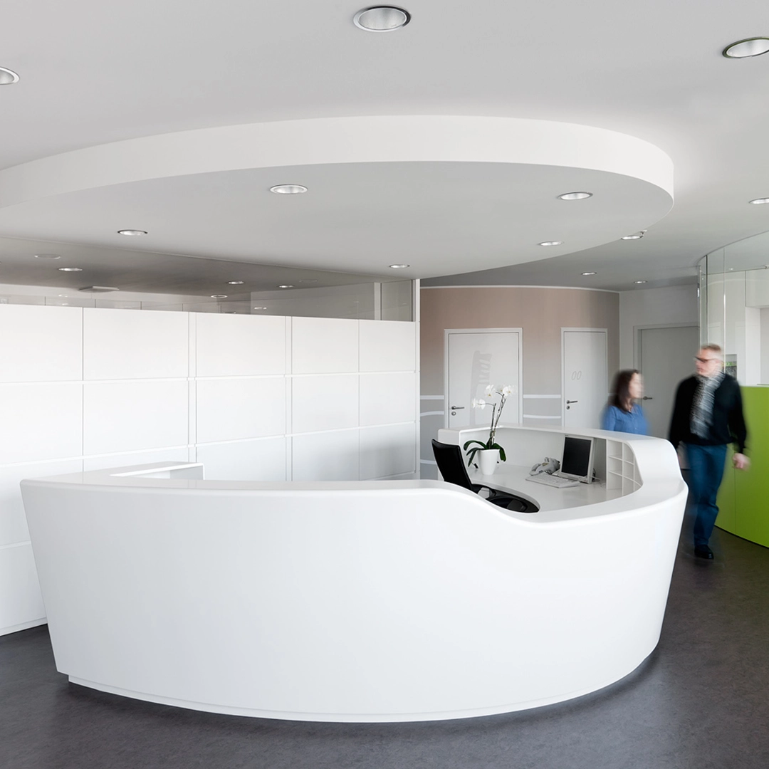 Modern white reception desk with curved design in a bright office lobby, featuring a computer, orchid plant, and two people walking nearby.