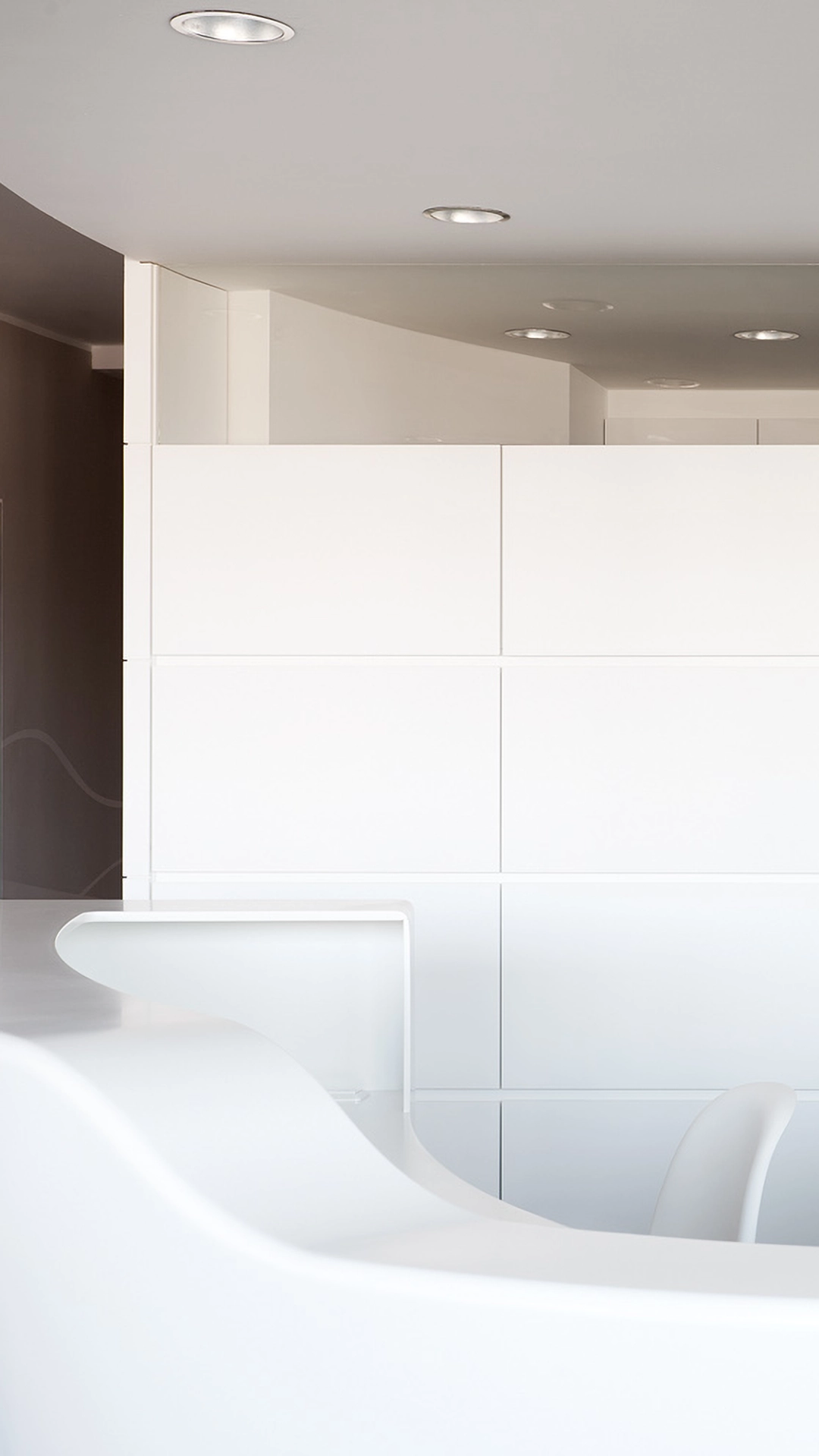 Modern kitchen with smooth white curved countertop and sleek white cabinetry under recessed ceiling lights.