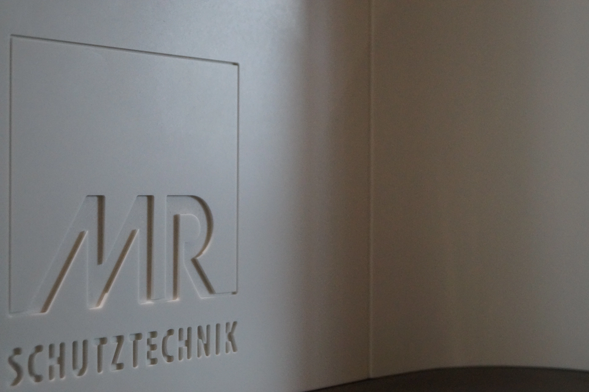 Embossed MR Schutztechnik logo on a smooth white surface with a soft shadow on the right.