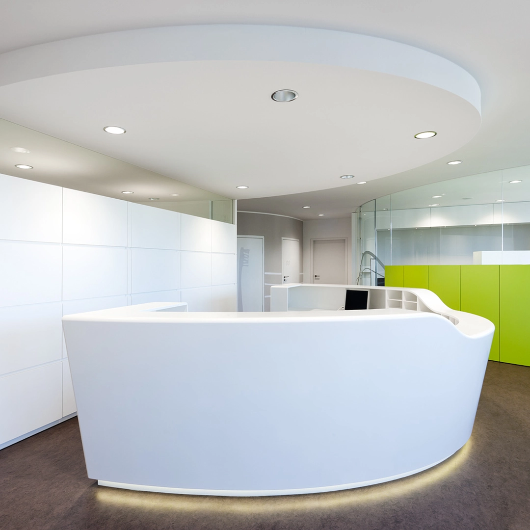 Modern white curved reception desk in a bright office with green cabinets and overhead recessed lighting.