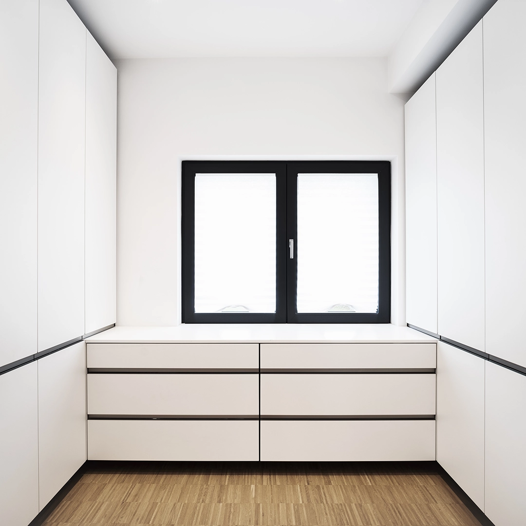 Minimalist white built-in cabinets and drawers under a black-framed window in a room with wooden flooring.