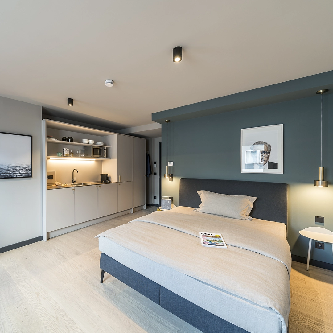 Modern studio bedroom with a gray upholstered bed, beige bedding, a kitchenette with cabinets, and minimalist decor on walls.