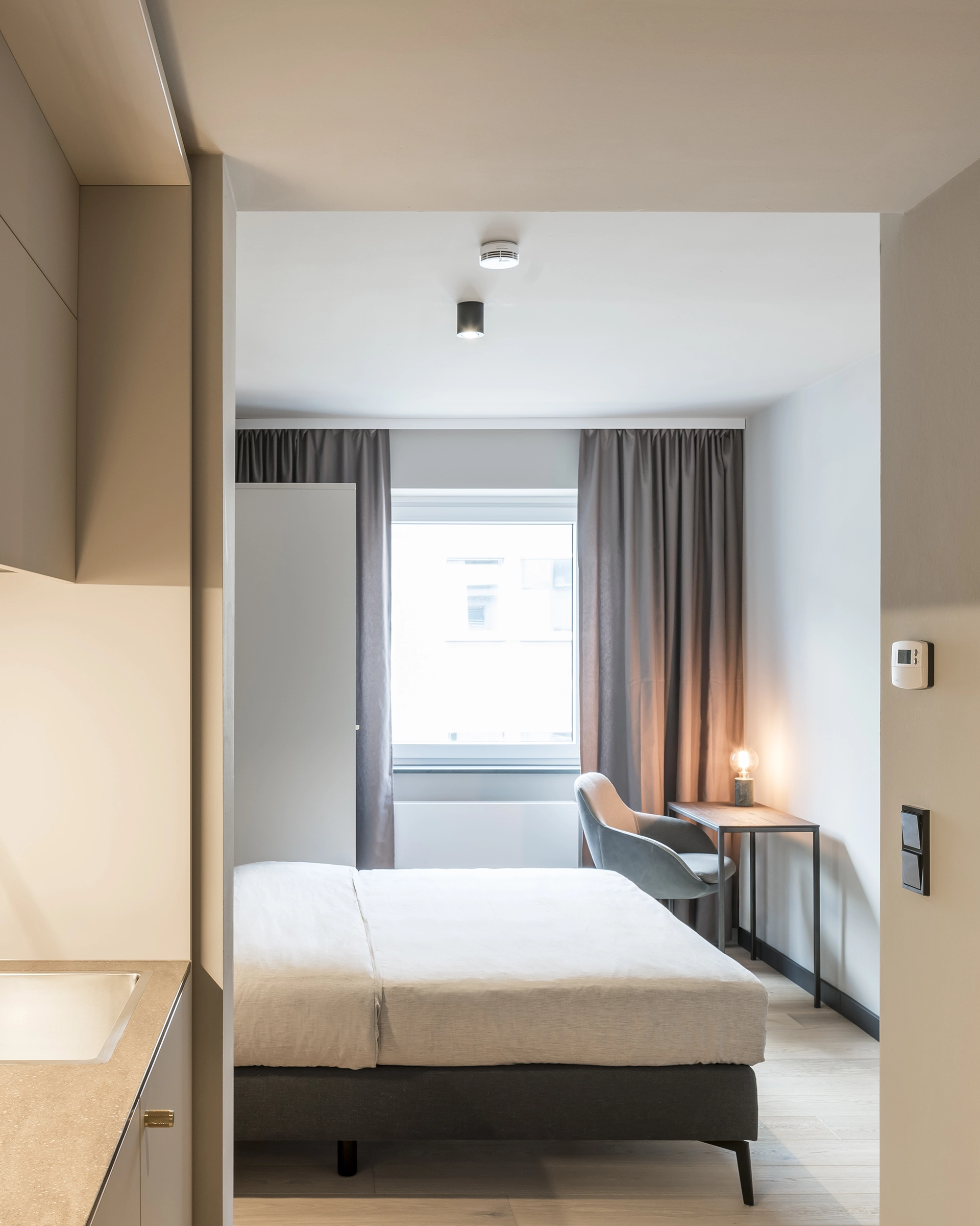 Minimalist bedroom with a bed, gray curtains, a window, a desk with a glowing lamp, and a blue chair.