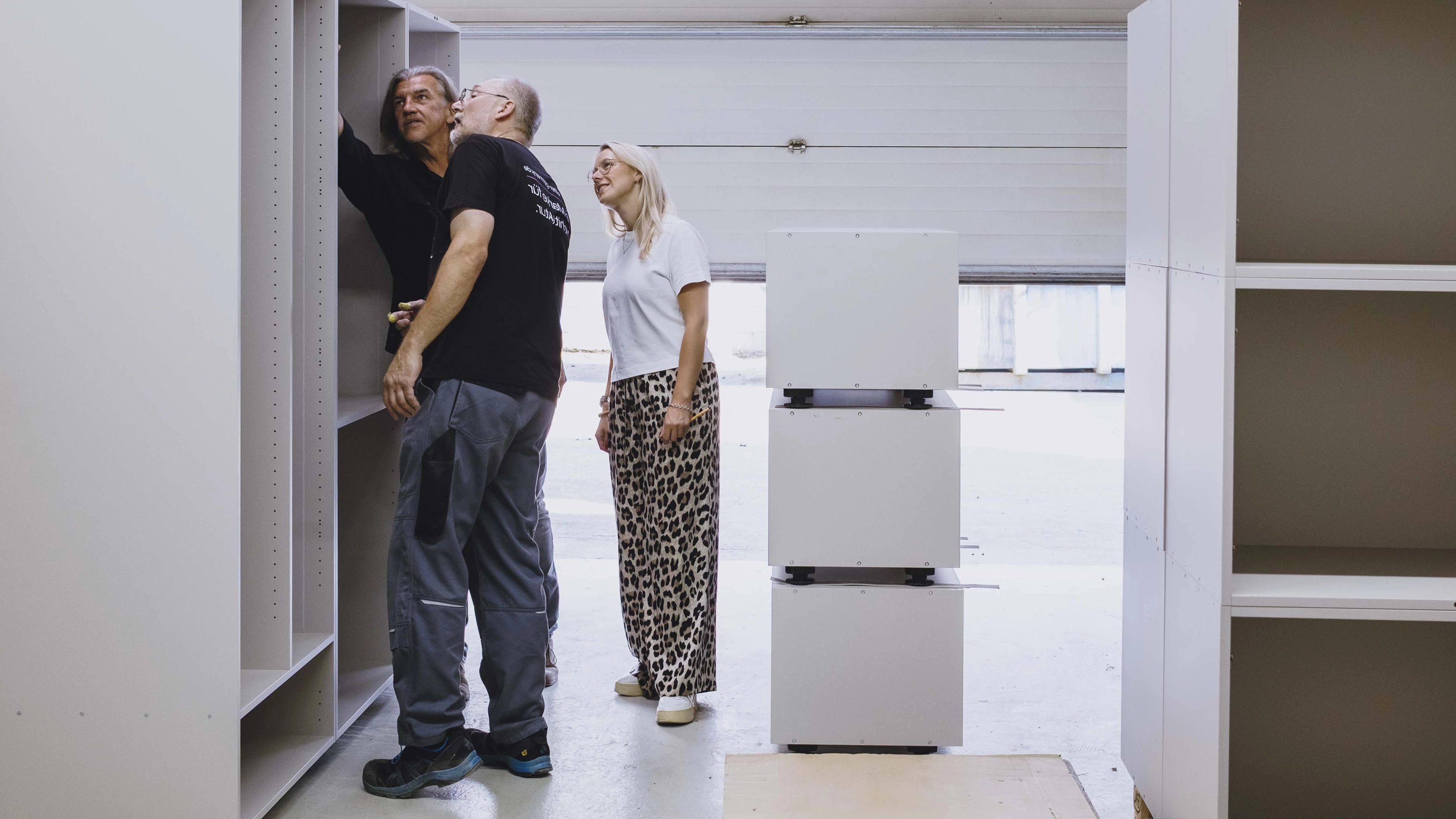 Three people examining large white modular shelving units in a spacious workshop.