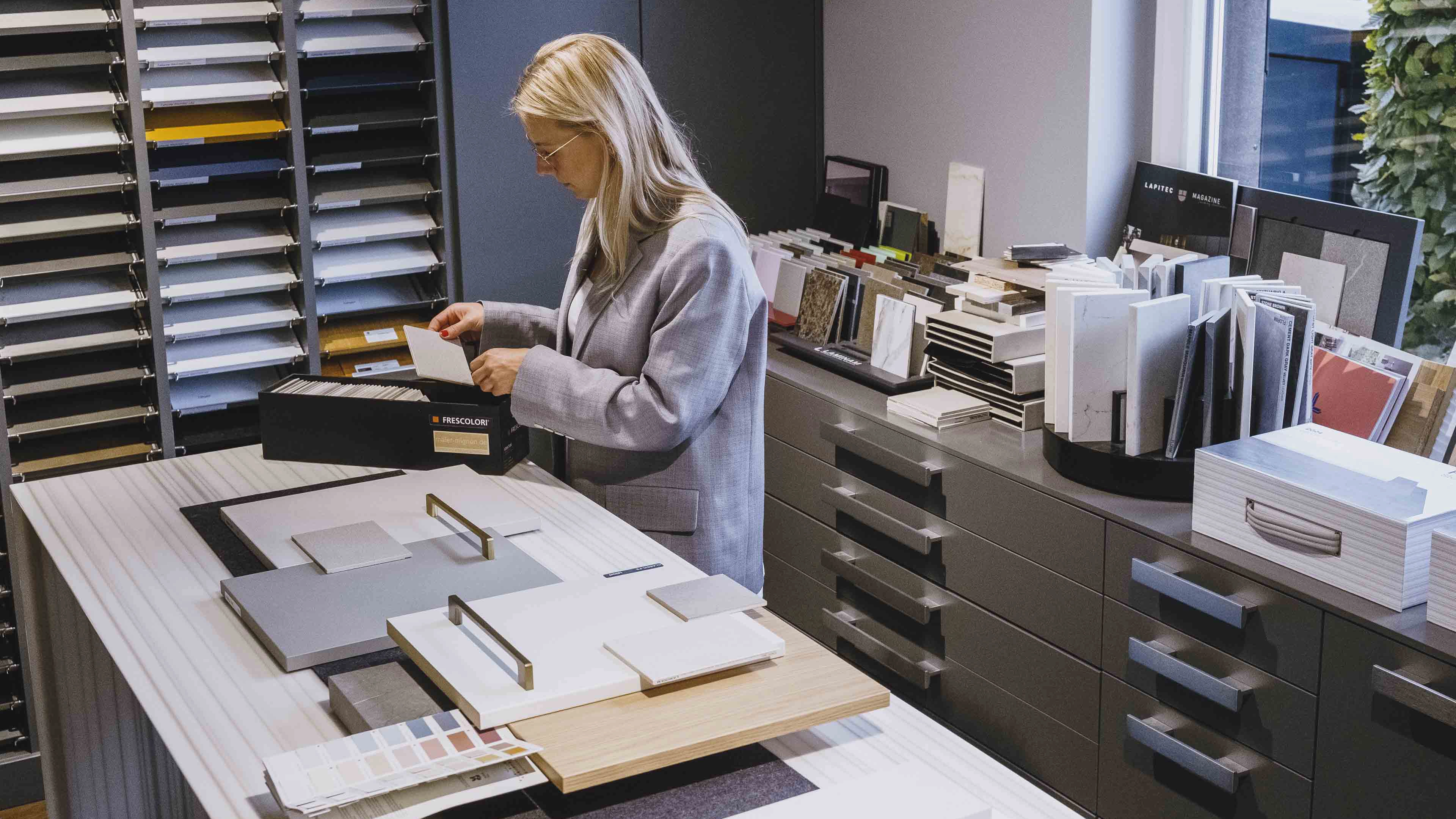 Woman examining sample tiles and color swatches in a modern showroom with cabinets and design materials.