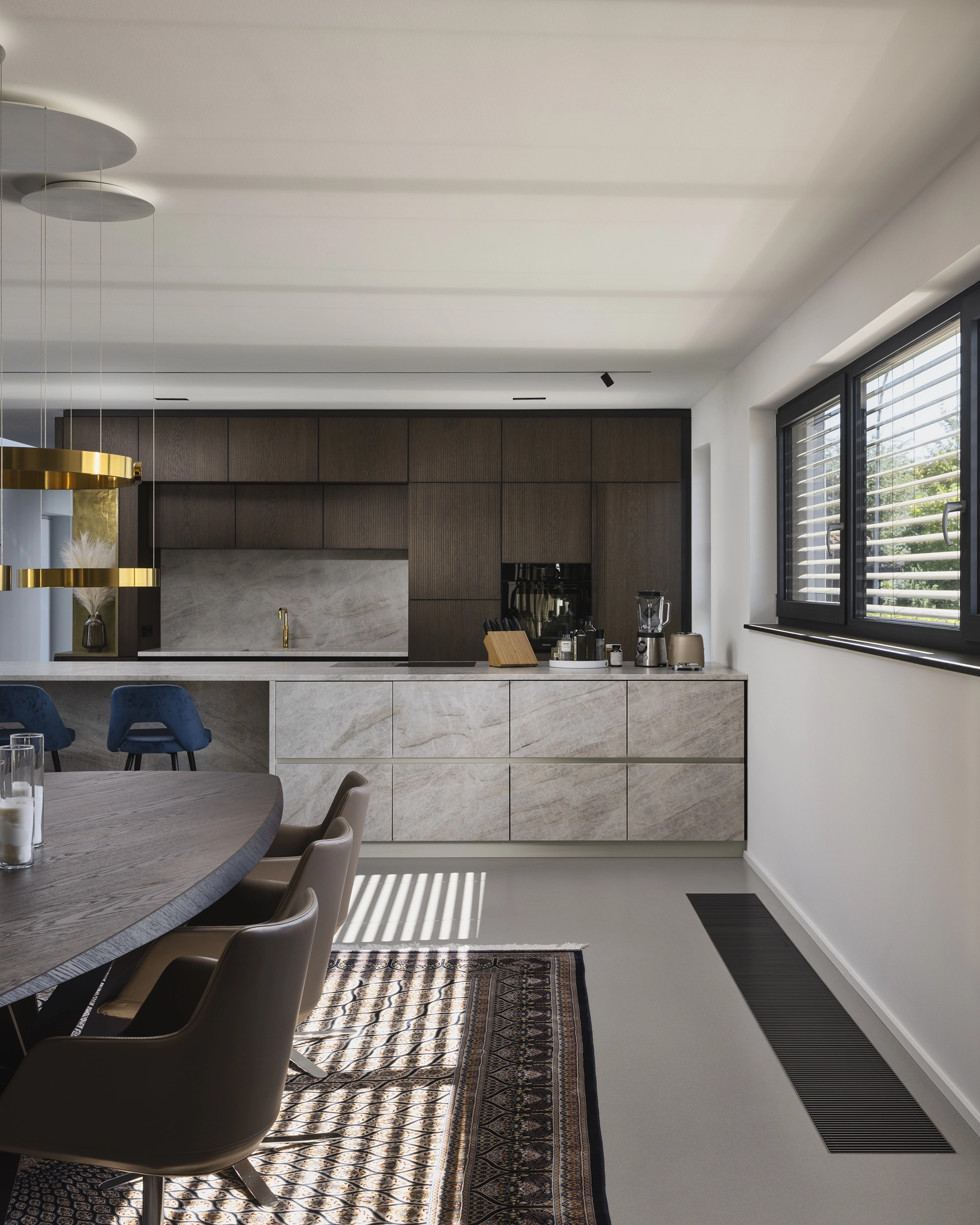 Modern kitchen with dark wood upper cabinets, light stone lower cabinets and countertop, blue bar stools, beige dining chairs, round wood table, and large window with blinds.