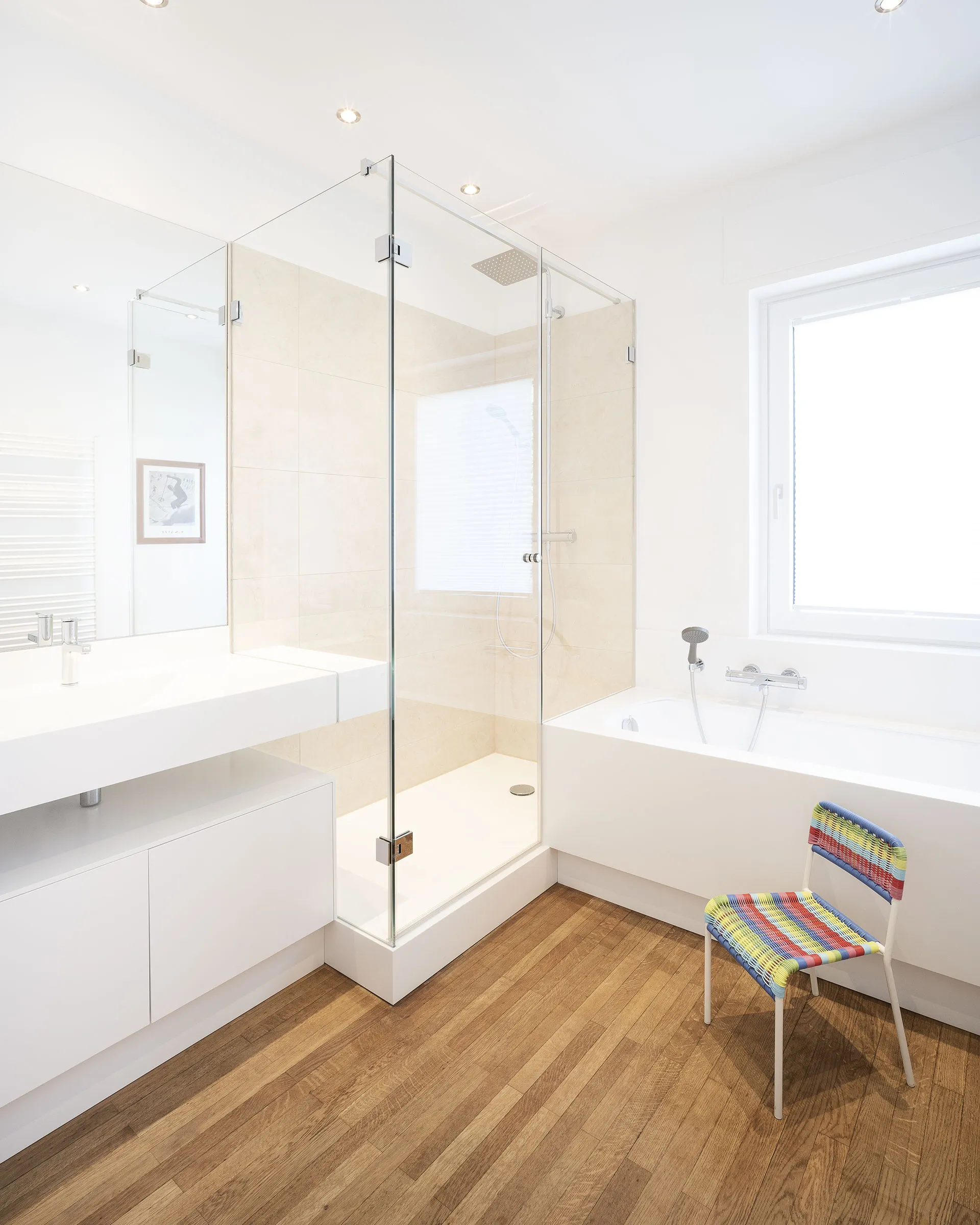 Bright modern bathroom with a glass-enclosed shower, white bathtub below a window, wooden floor, and a colorful woven chair.