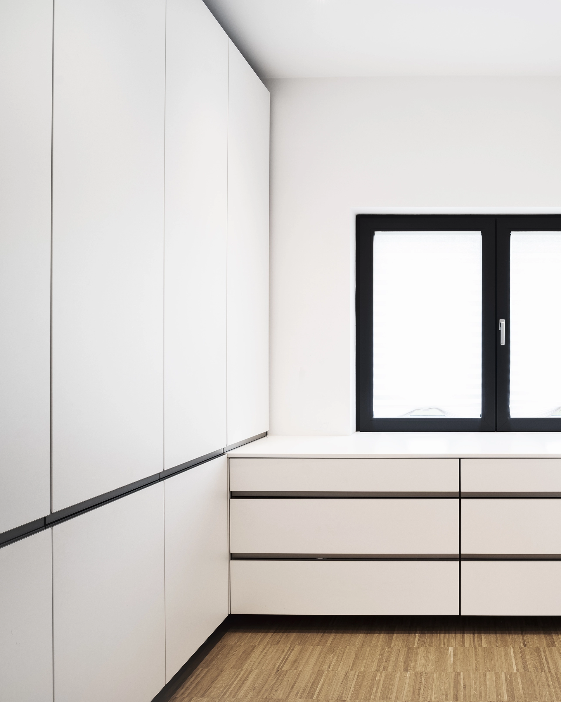 Corner of a modern white kitchen with sleek cabinets and drawers under a window with black frame.