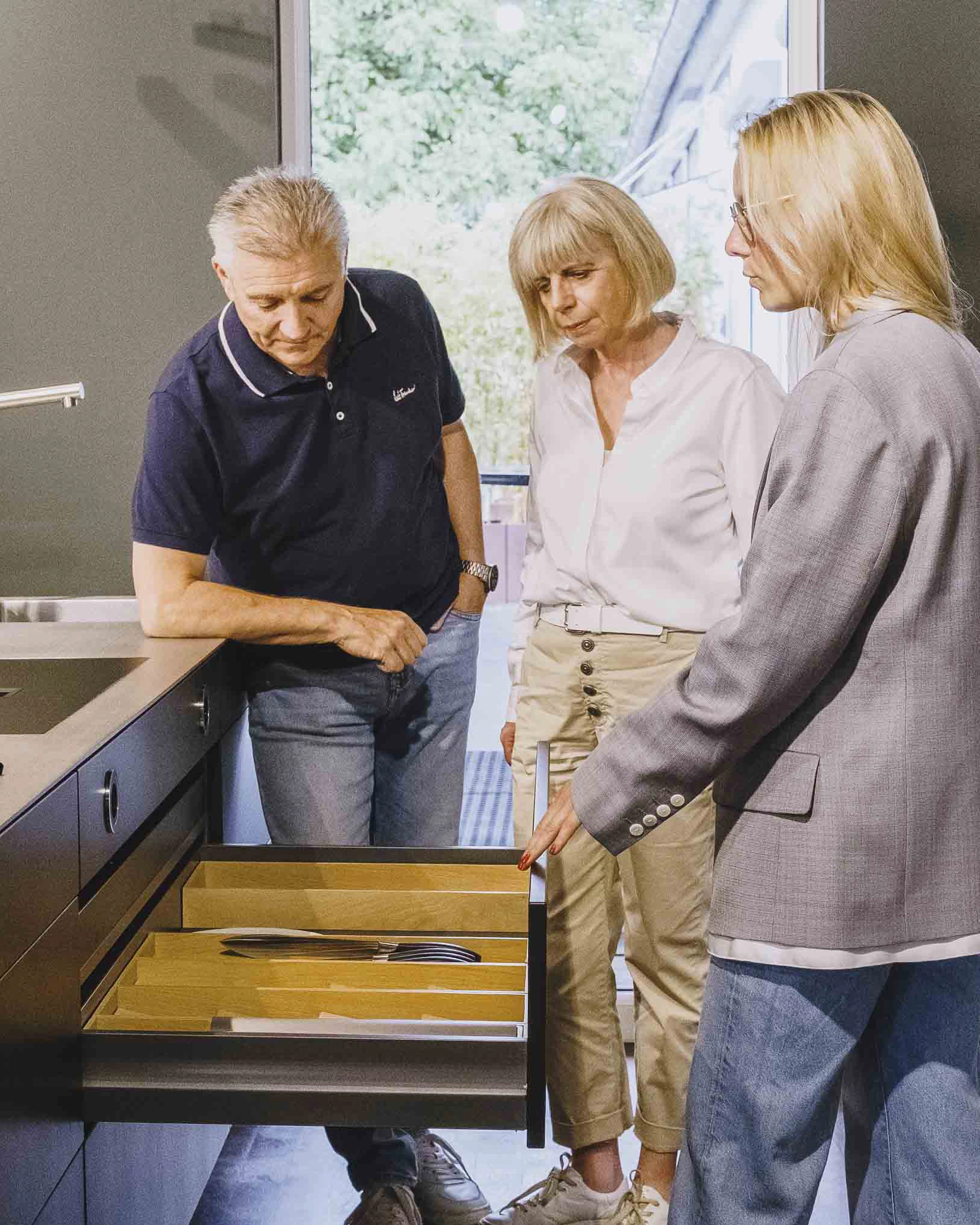 Three people inspecting an open kitchen drawer with organized wooden cutlery trays.