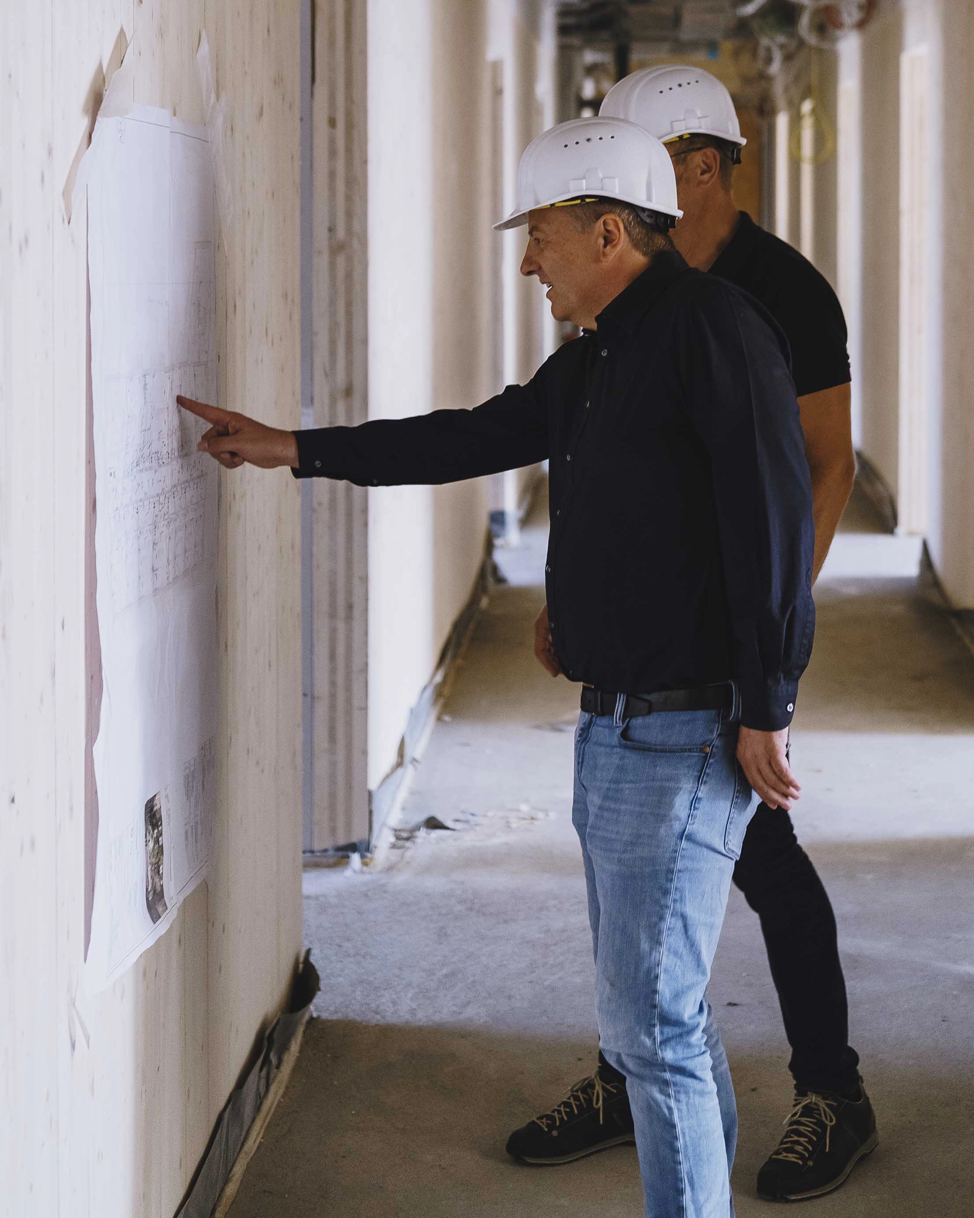 Two men wearing white construction helmets reviewing a blueprint pinned to a wall inside a building under construction.