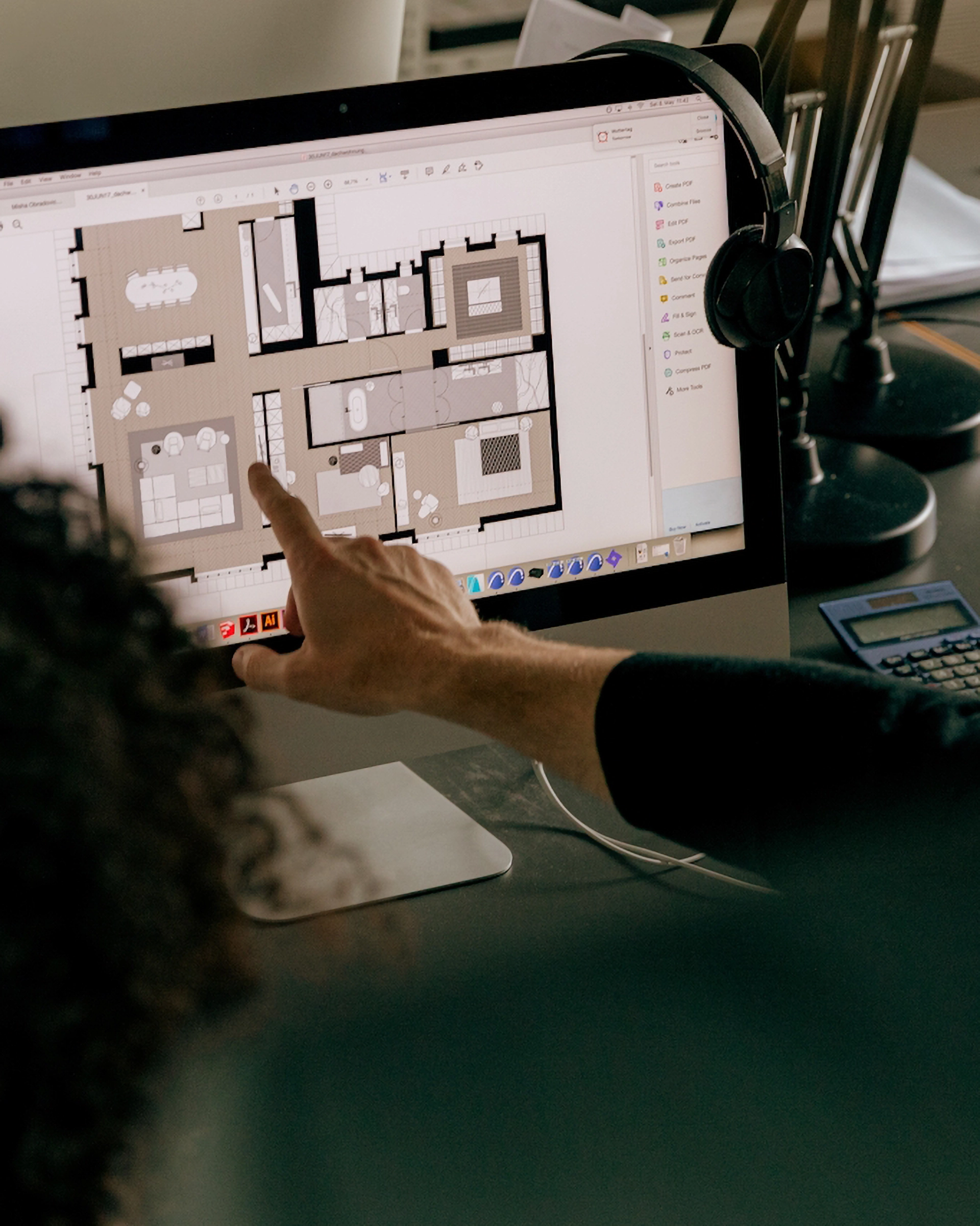 Person pointing at a computer screen displaying a digital floor plan layout, with headphones hanging on the monitor and a calculator on the desk.