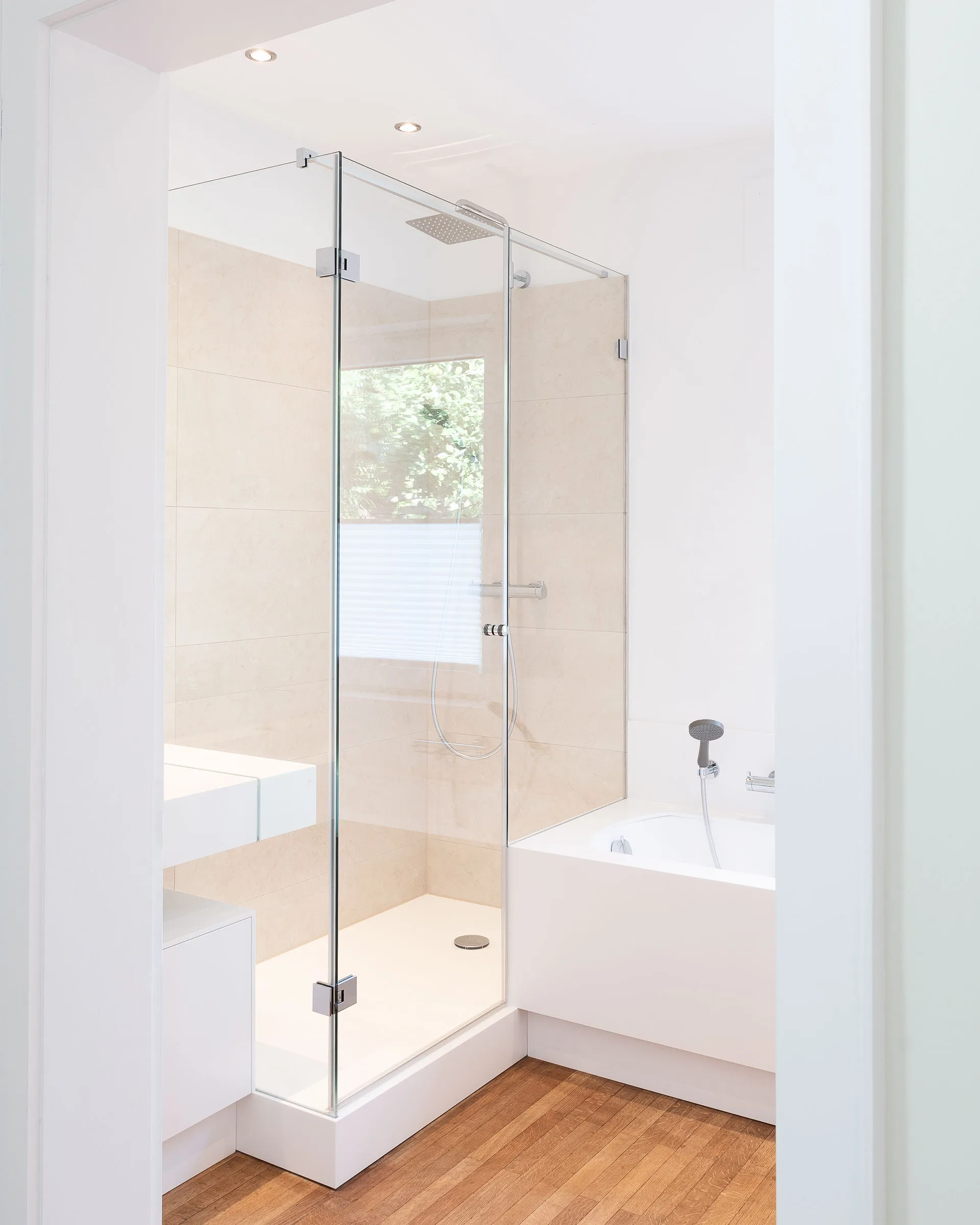 Minimalist bathroom with a glass-enclosed shower, beige tiles, white bathtub, and wooden floor.