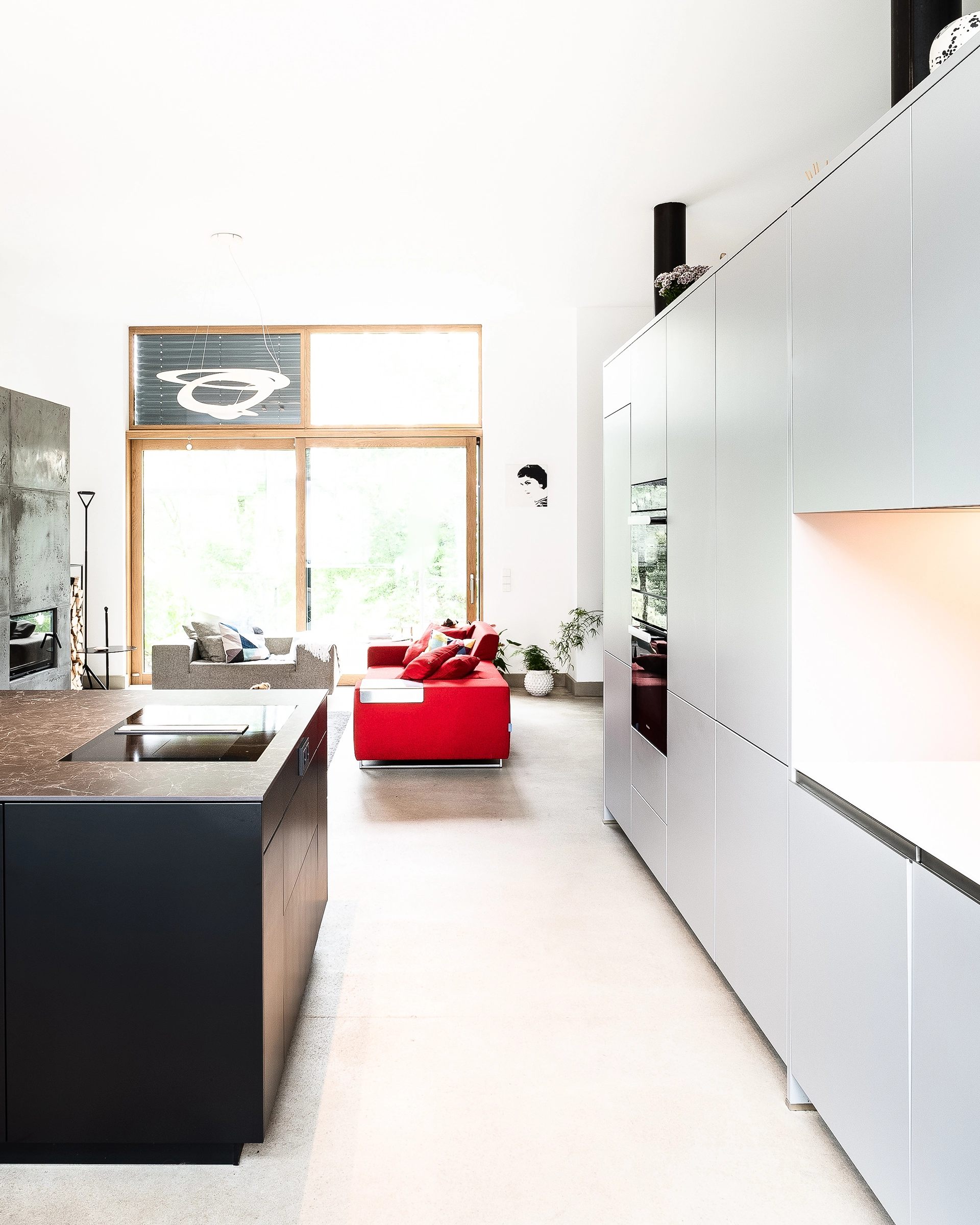 Bright modern kitchen with black island, white cabinets, and red and gray sofas near large wooden-framed window.