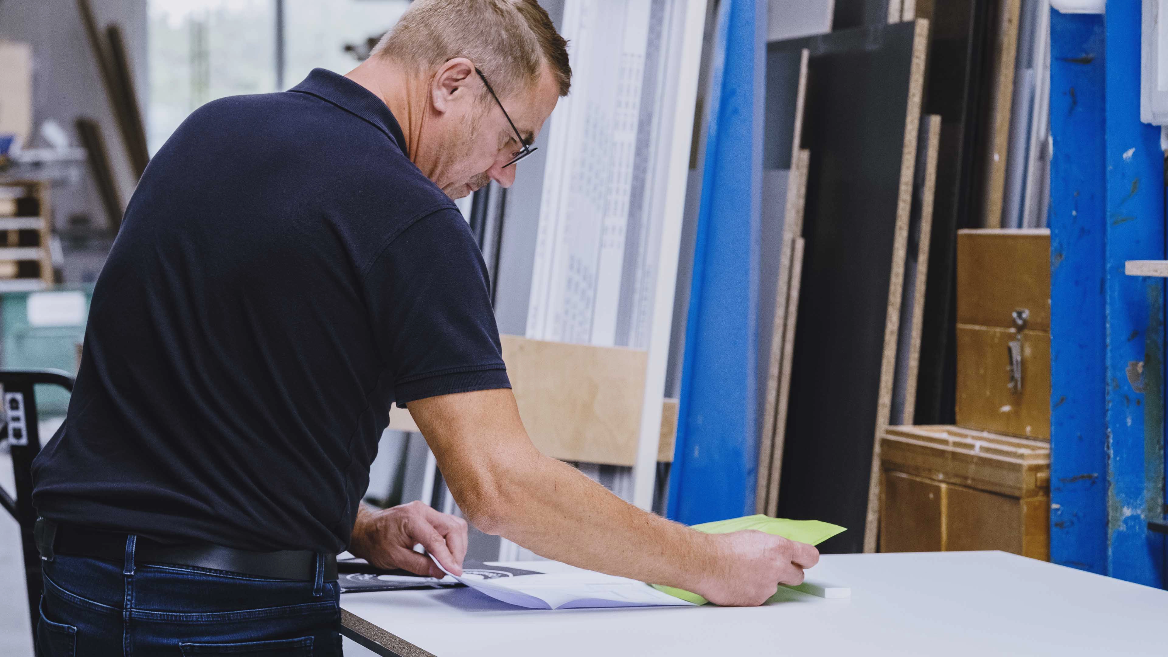 Man in a black polo shirt reading papers at a workshop table with blue and wooden materials in the background.