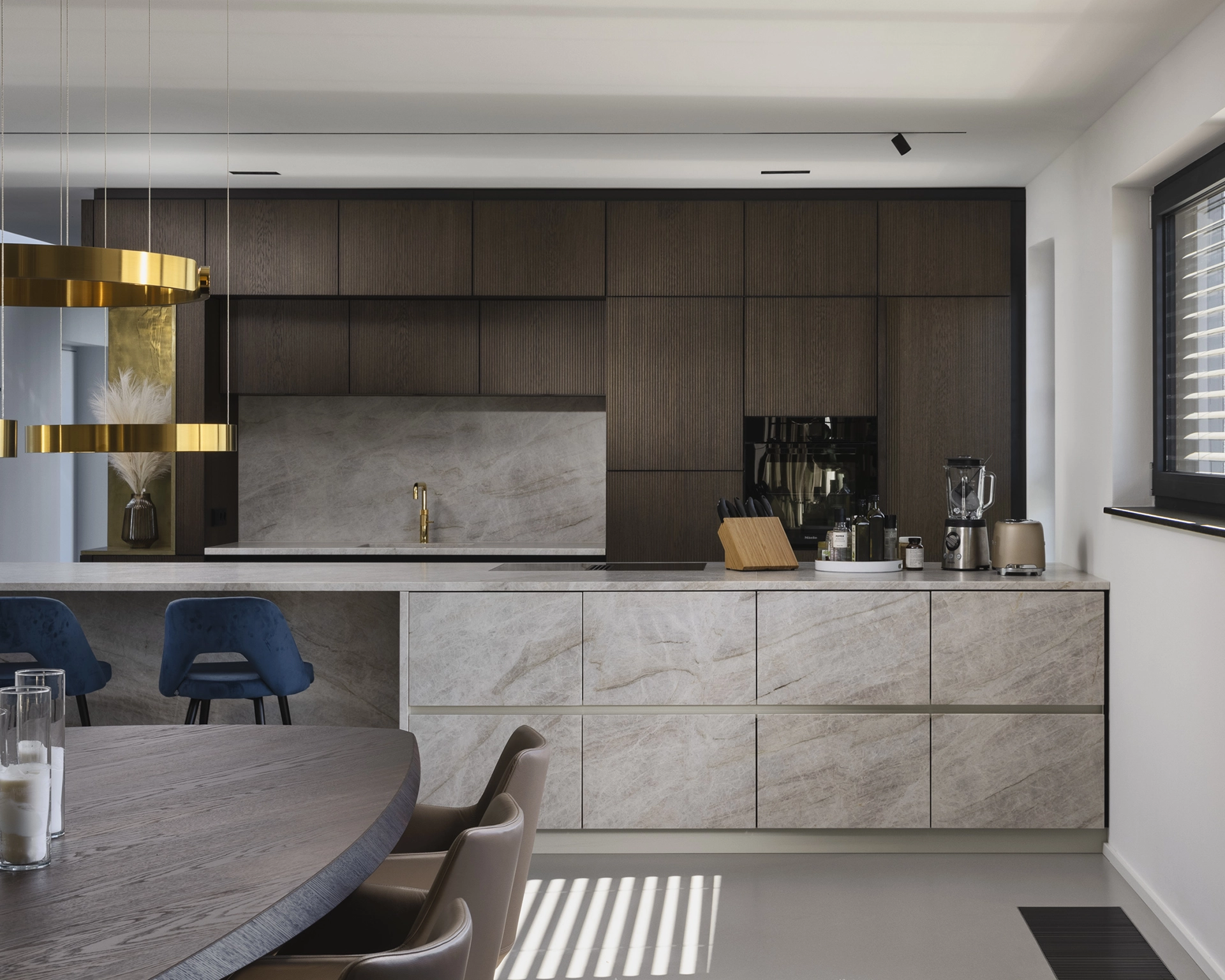 Modern kitchen with dark wood upper cabinets, marble-effect lower cabinets and countertop, blue bar stools, gold pendant lights, and a dining table with brown chairs.