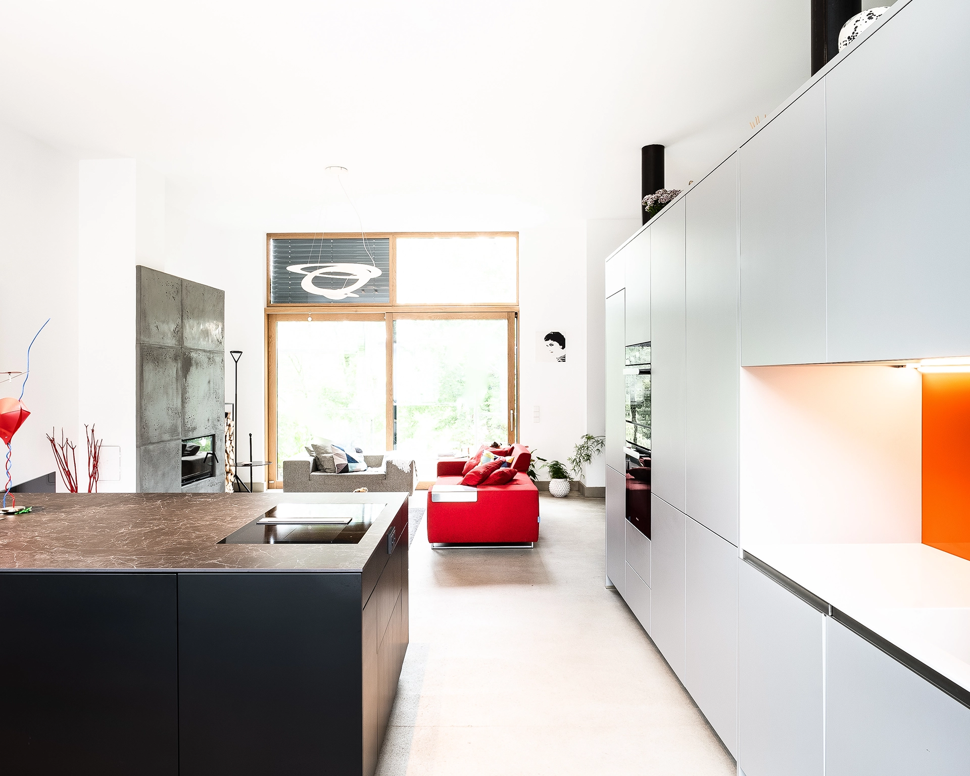 Bright open-plan kitchen and living area with gray and red sofas, modern black kitchen island, white cabinets, and large windows.