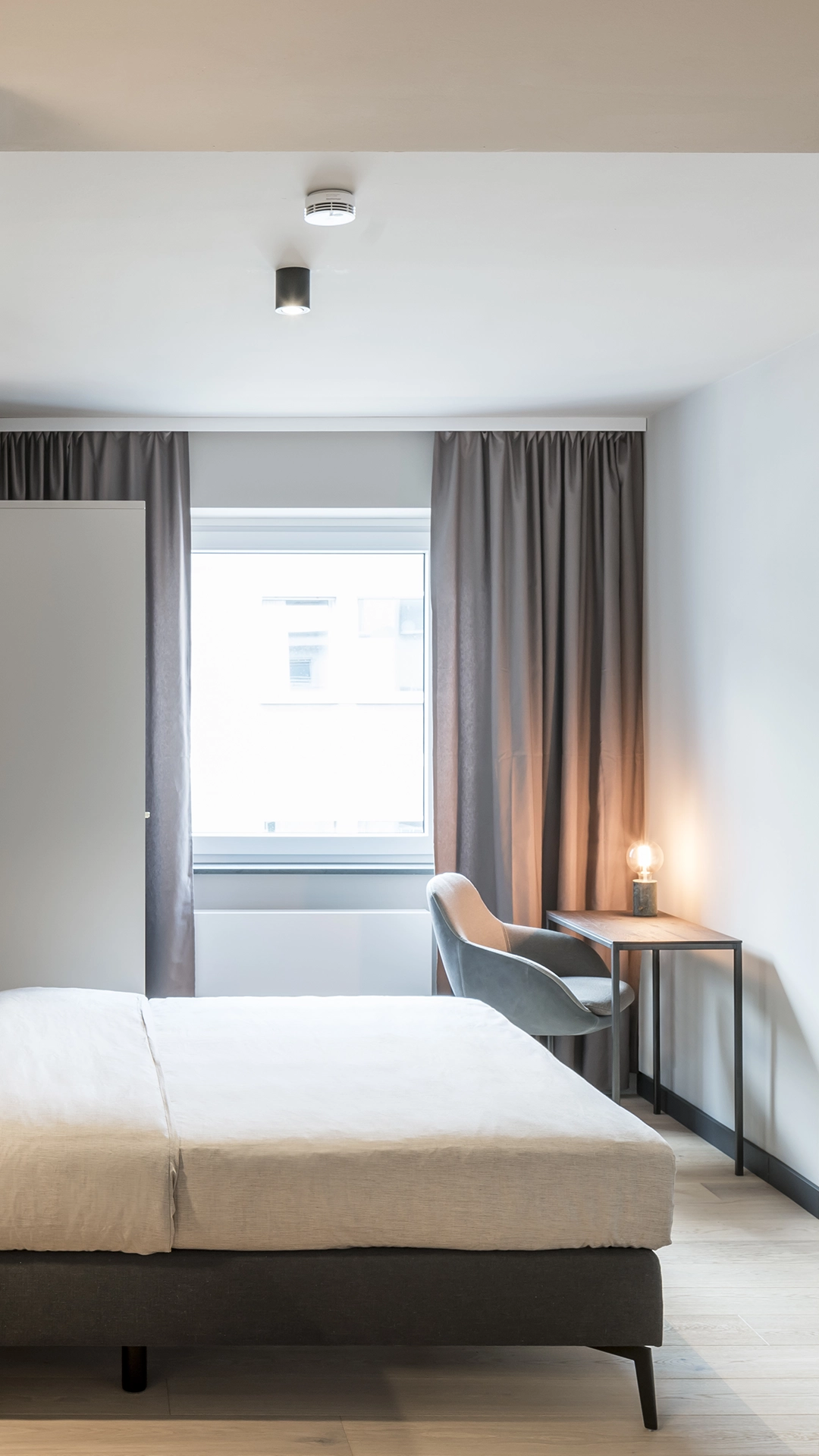 Minimalist bedroom with a neatly made bed, gray curtains framing a window, a gray chair, and a small wooden table with a glowing bulb lamp.