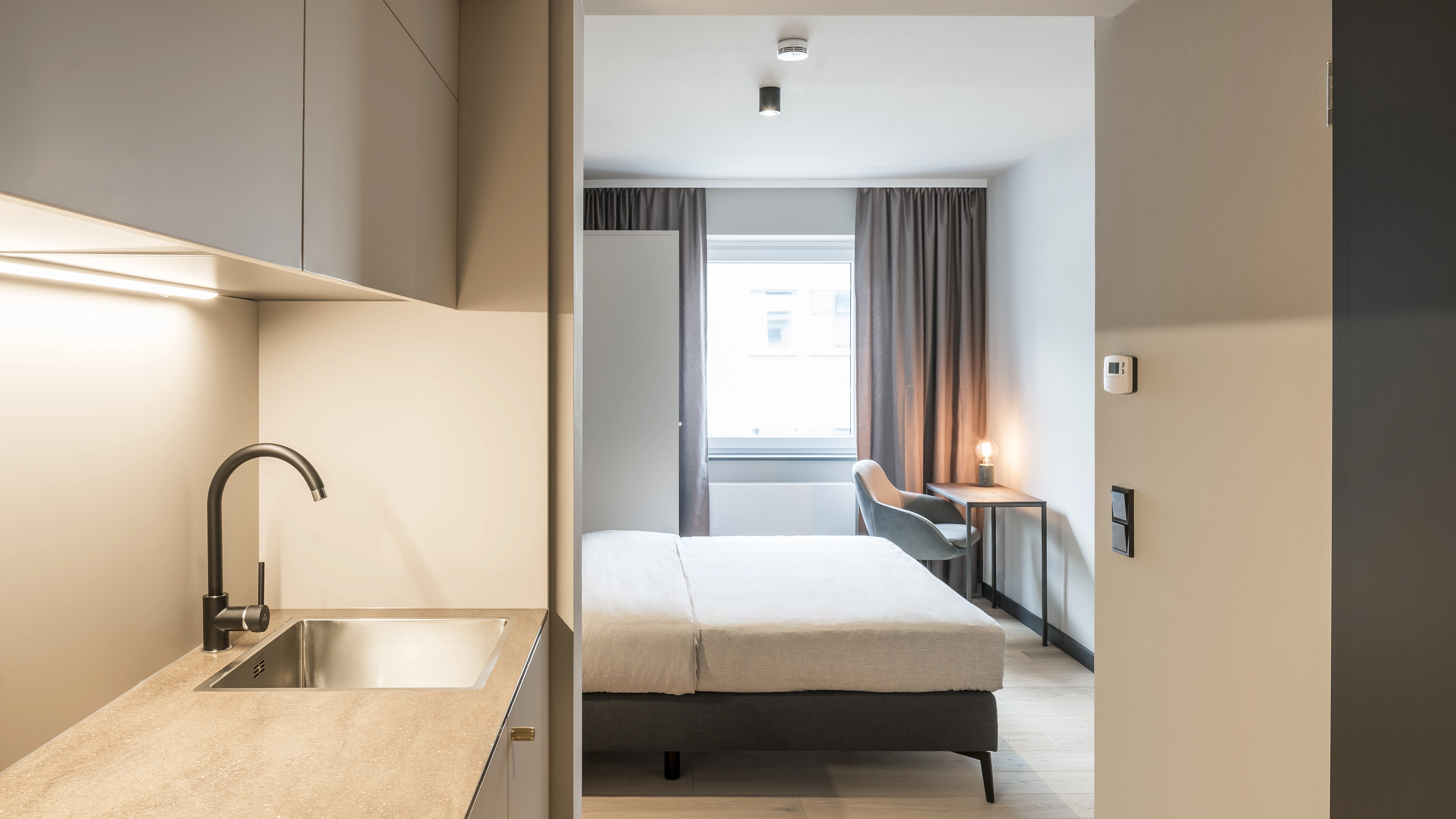 Minimalist studio apartment with a kitchenette, bed, desk with lamp, and window with curtains.