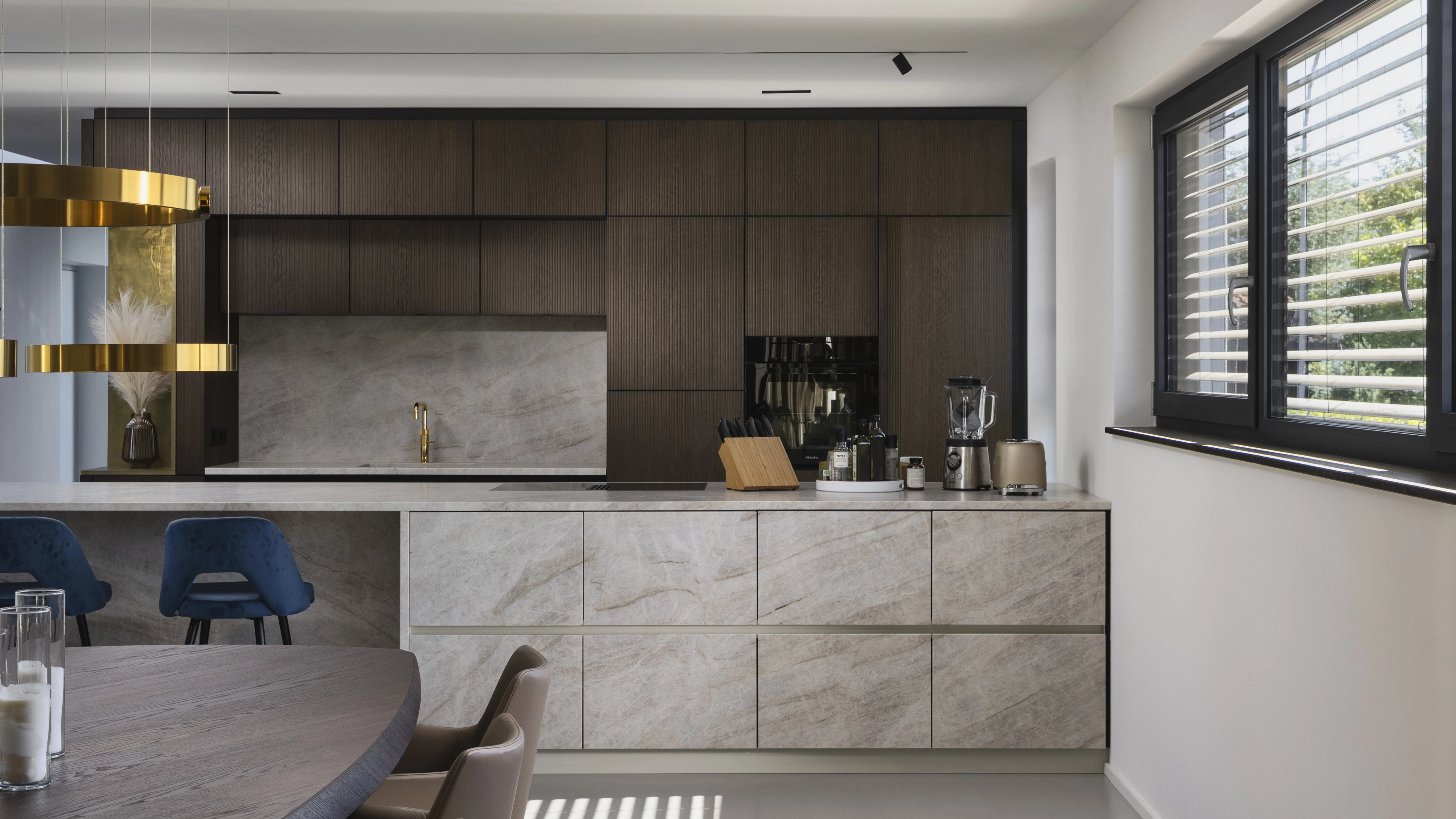 Modern kitchen with dark wood upper cabinets, light marble lower cabinets, gold faucet, blue bar stools, and large windows with blinds.