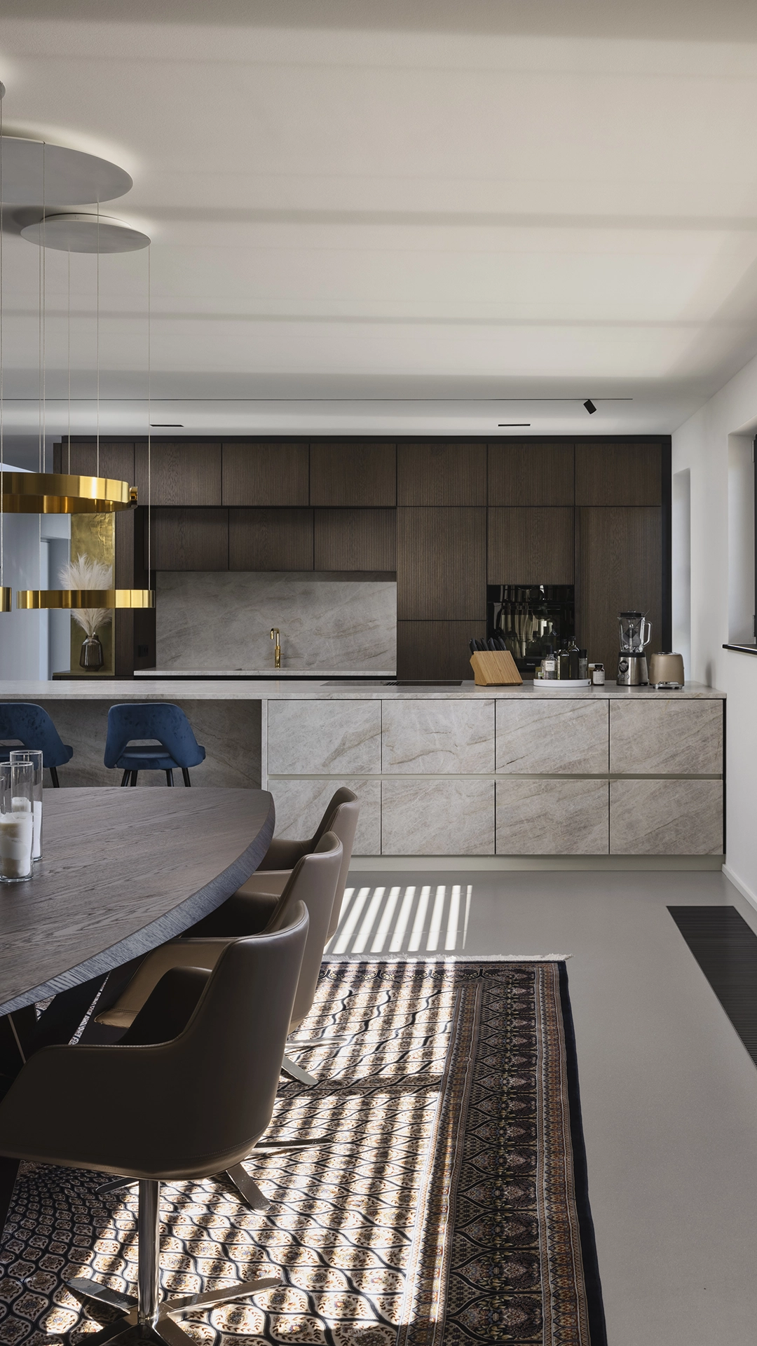 Modern kitchen with dark wood cabinets, marble countertops, gold faucet, blue bar stools, and a round dining table with brown chairs on a patterned rug.