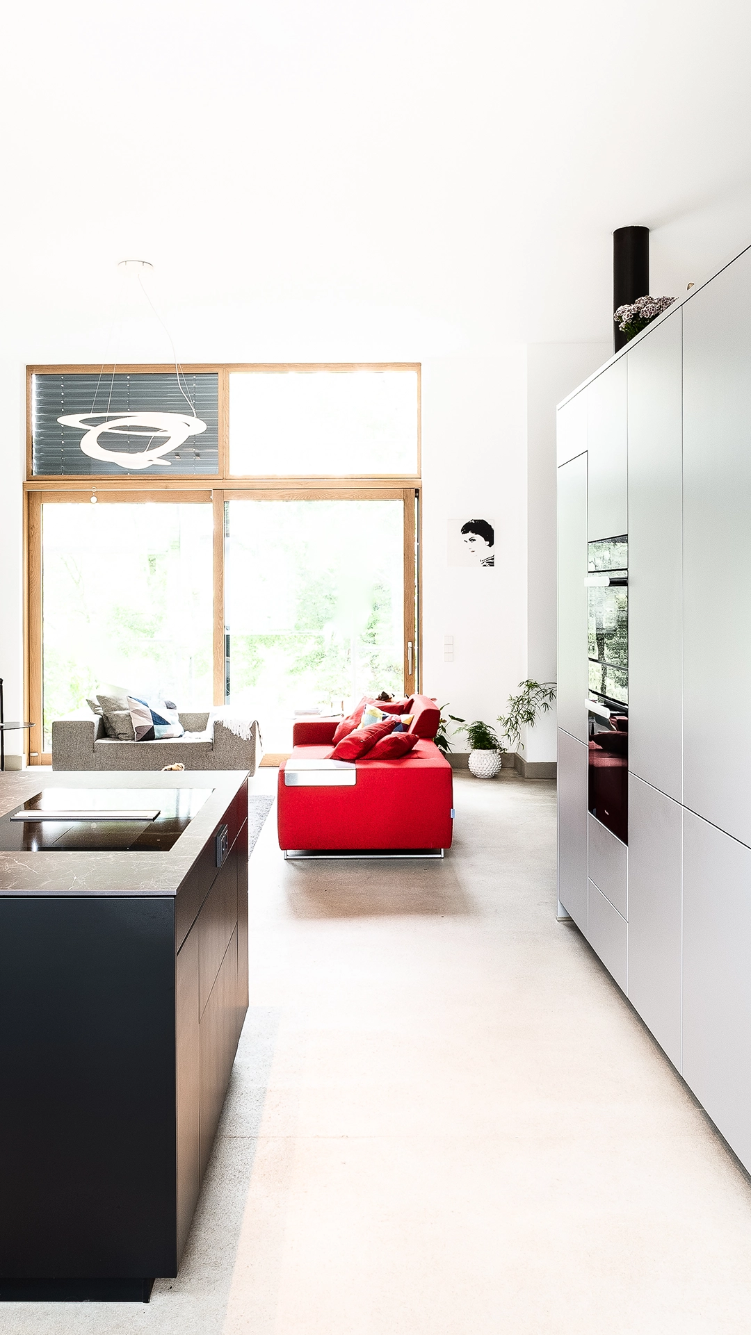 Bright modern living space with a red sofa, gray armchair, black kitchen island, and large windows with wooden frames.