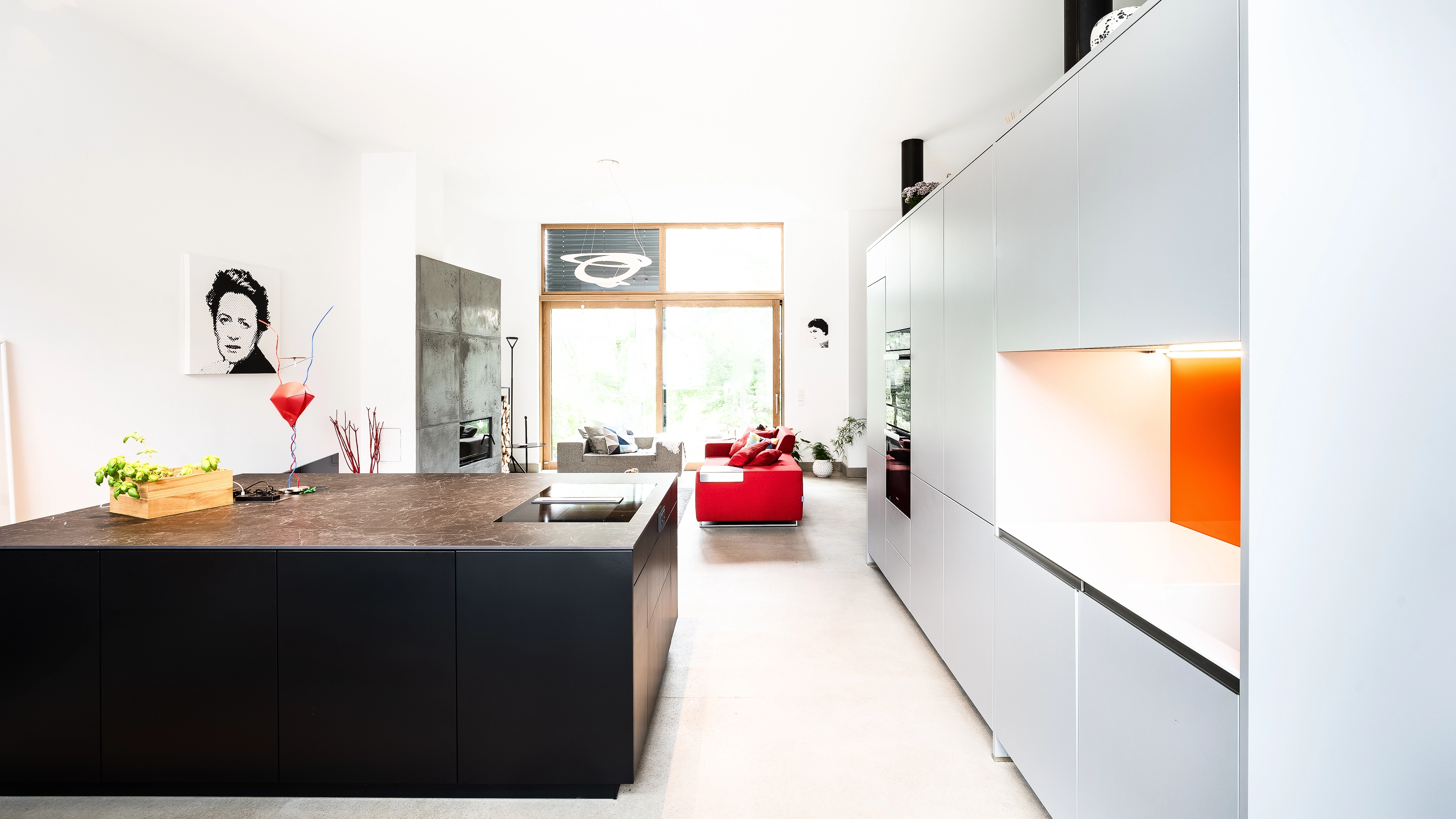 Bright modern open-plan living space with black kitchen island, white cabinets with orange accent, and red and gray sofas near large window.