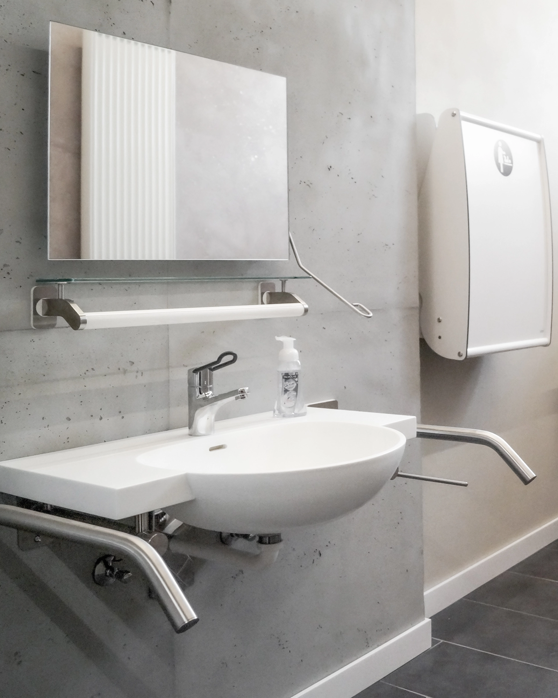 Barrierefreies weißes wandmontiertes Waschbecken mit Spiegel, Handlauf und Seifenspender in einem sanitärbereich.