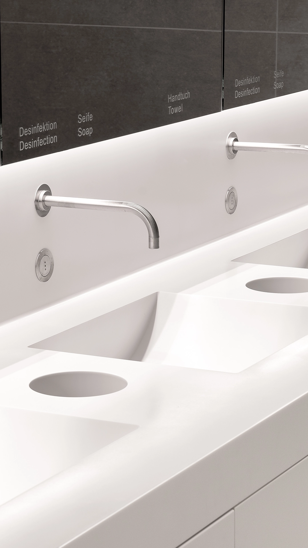 Modern white bathroom sink with two wall-mounted stainless steel faucets and mirrored cabinets labeled in German and English for disinfection, soap, and towel.