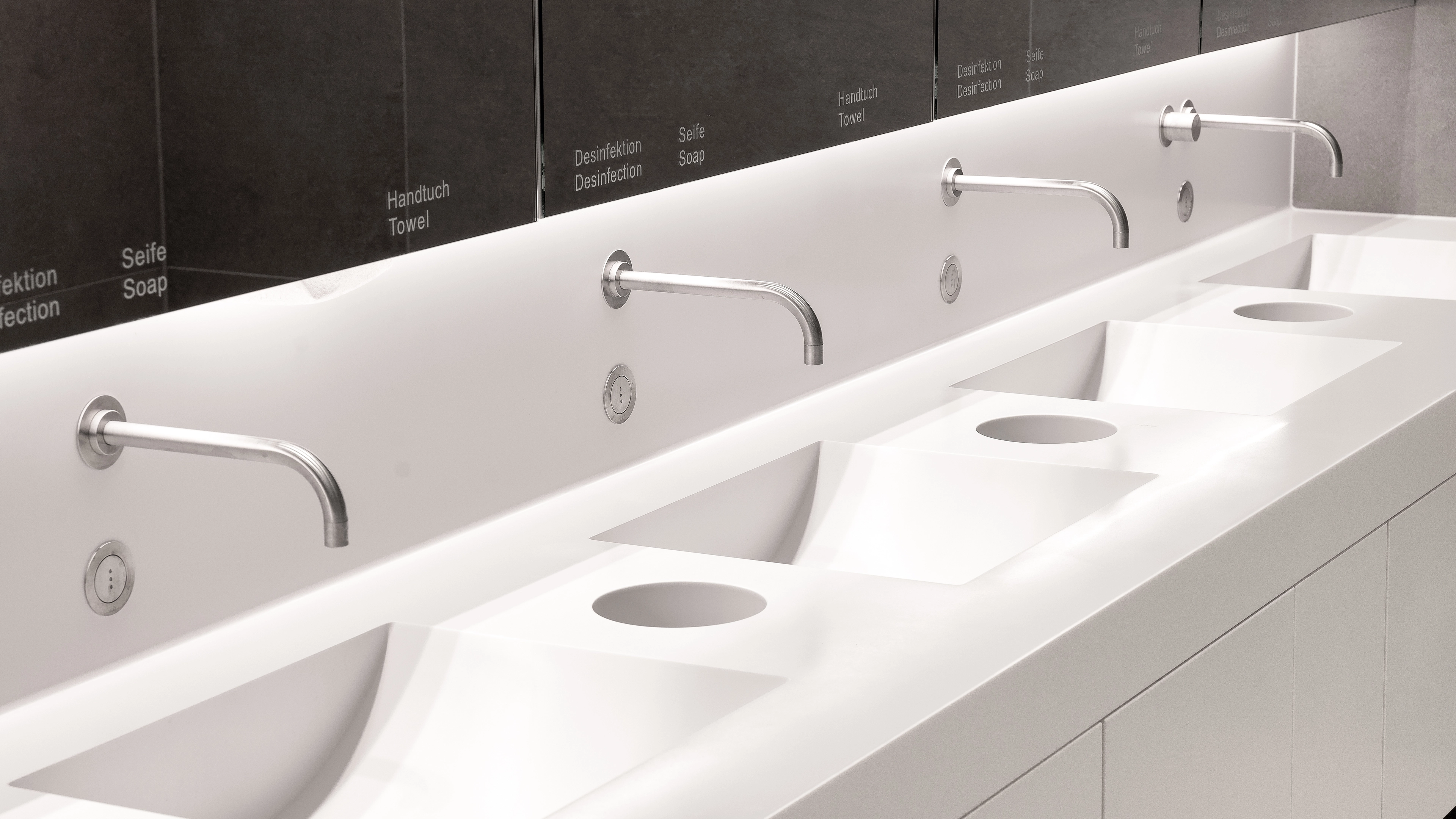 Modern white bathroom sink countertop with three streamlined metal faucets mounted on the wall and a large mirror with English and German labels for towel, soap, and disinfection.
