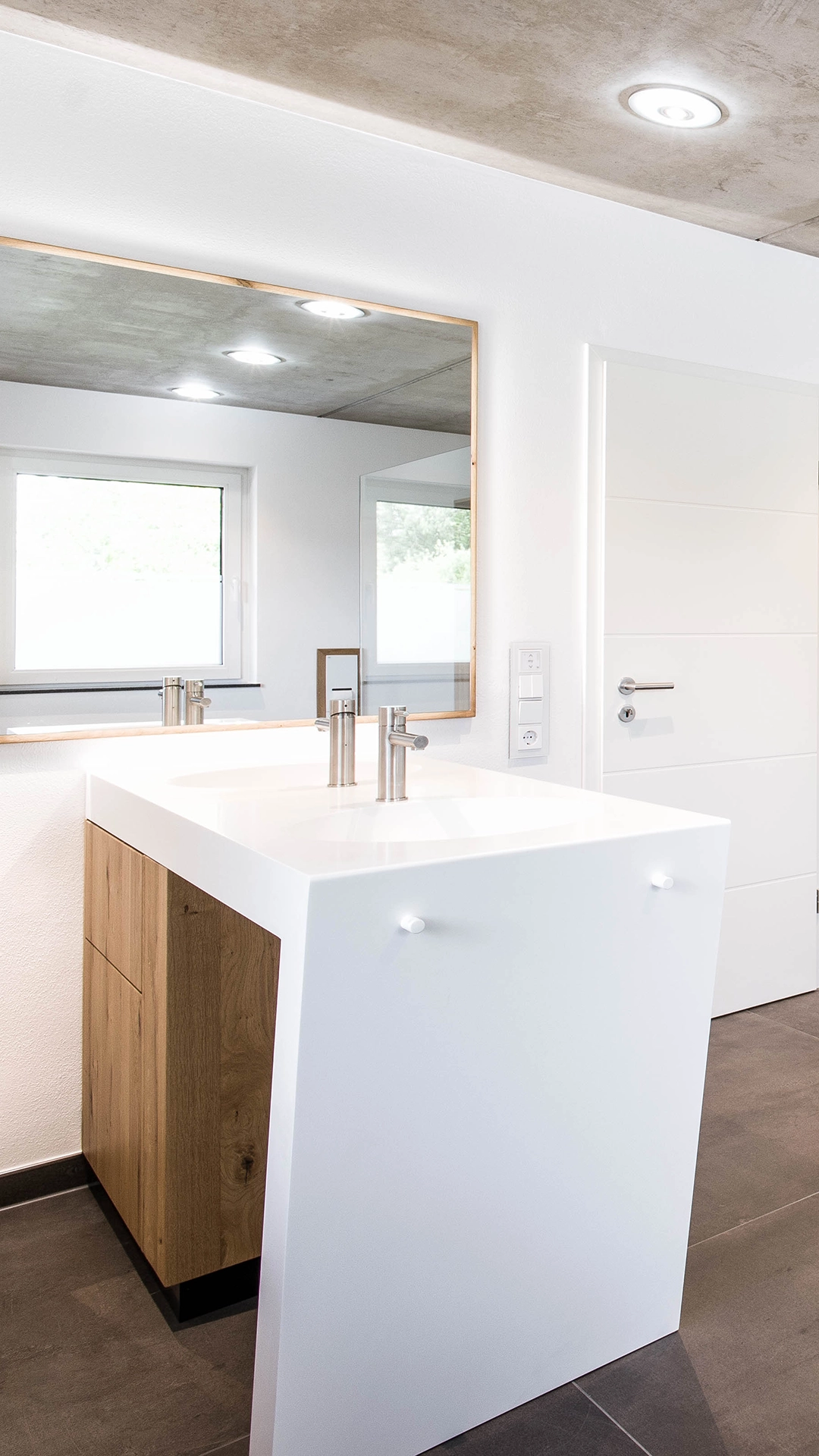 Modern bathroom sink with wooden cabinet, white countertop, and large mirror reflecting ceiling lights and a window.