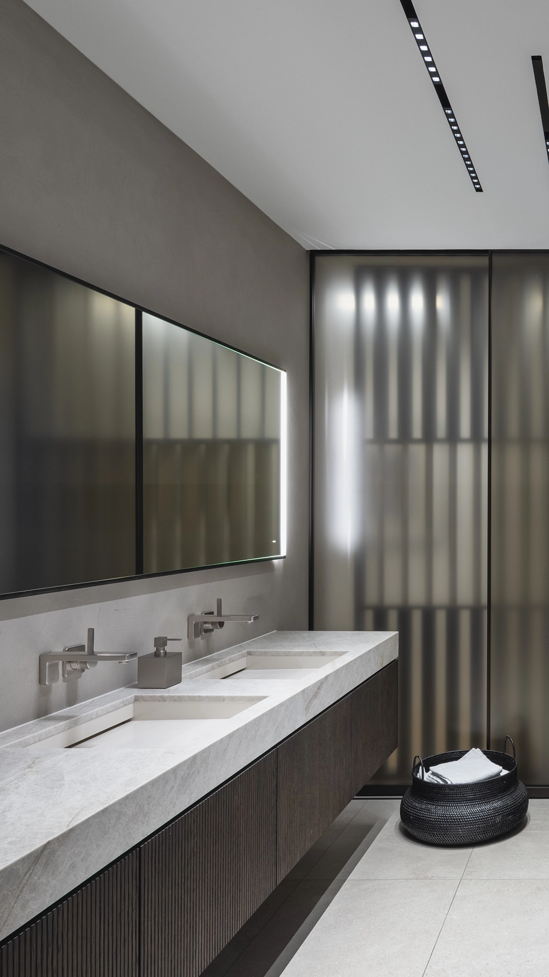 Modern bathroom with double sinks set in a white marble countertop, wall-mounted faucets, large illuminated mirror, frosted glass partition, and black basket with white towels on tiled floor.