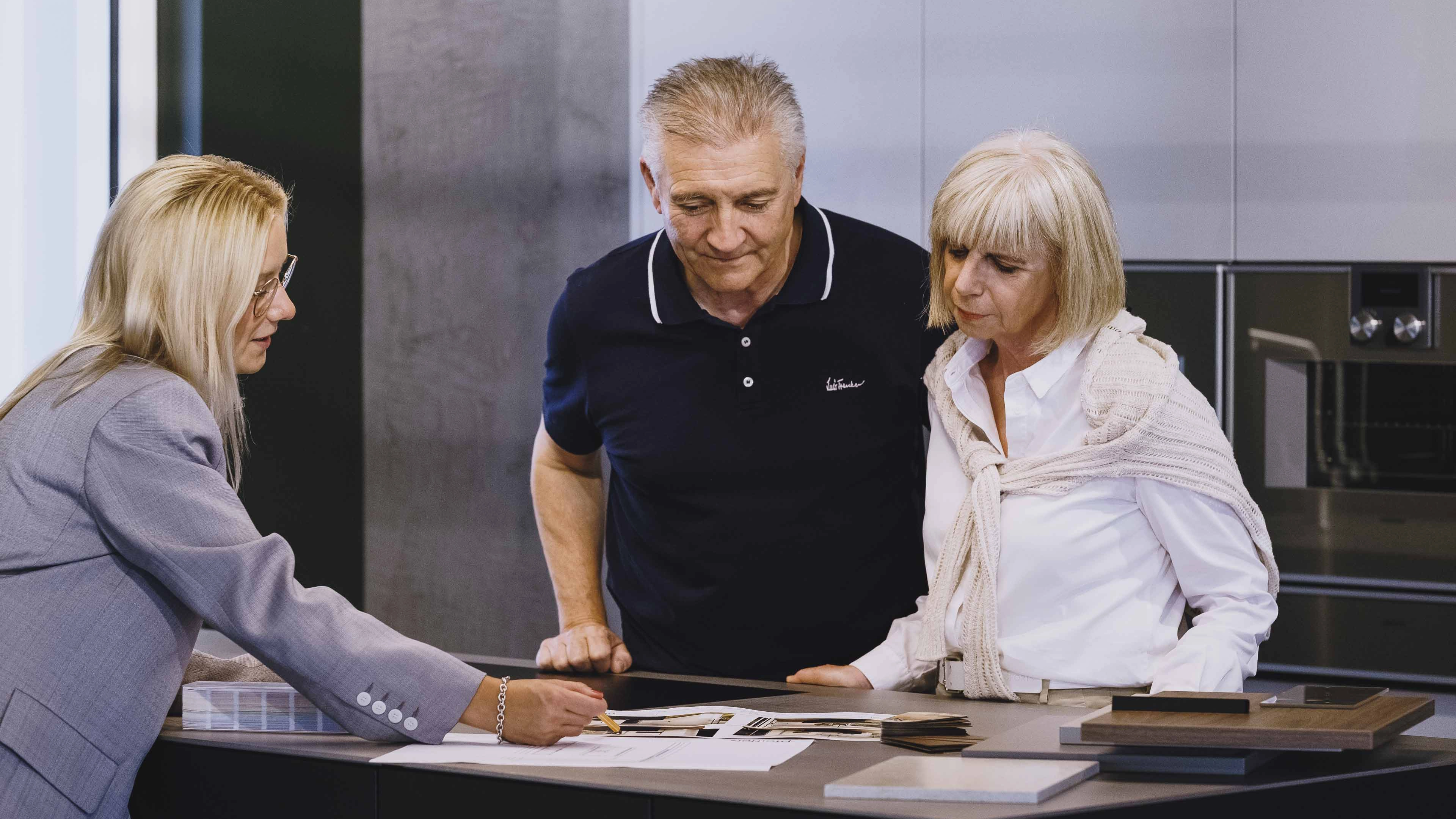 Three adults reviewing design plans and material samples on a kitchen counter.