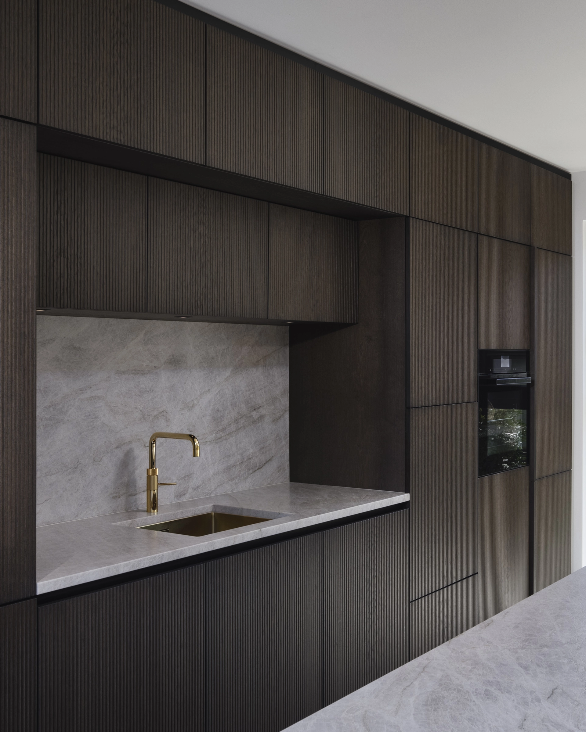 Modern kitchen with dark wood textured cabinets, a grey marble countertop and backsplash, and a gold faucet over a built-in sink.