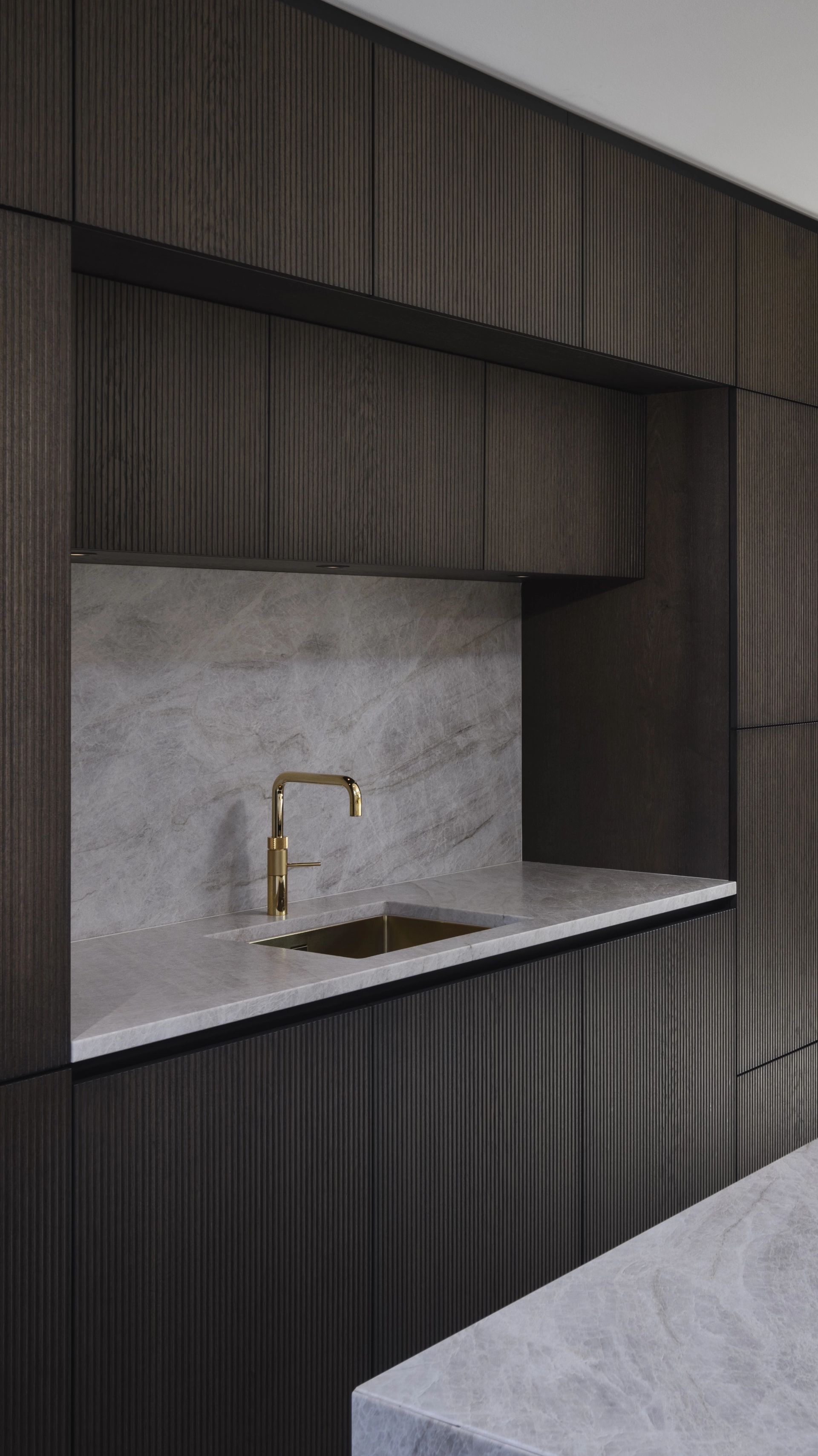 Modern kitchen with dark ribbed cabinetry, a gray marble countertop and backsplash, and a gold faucet above a built-in sink.