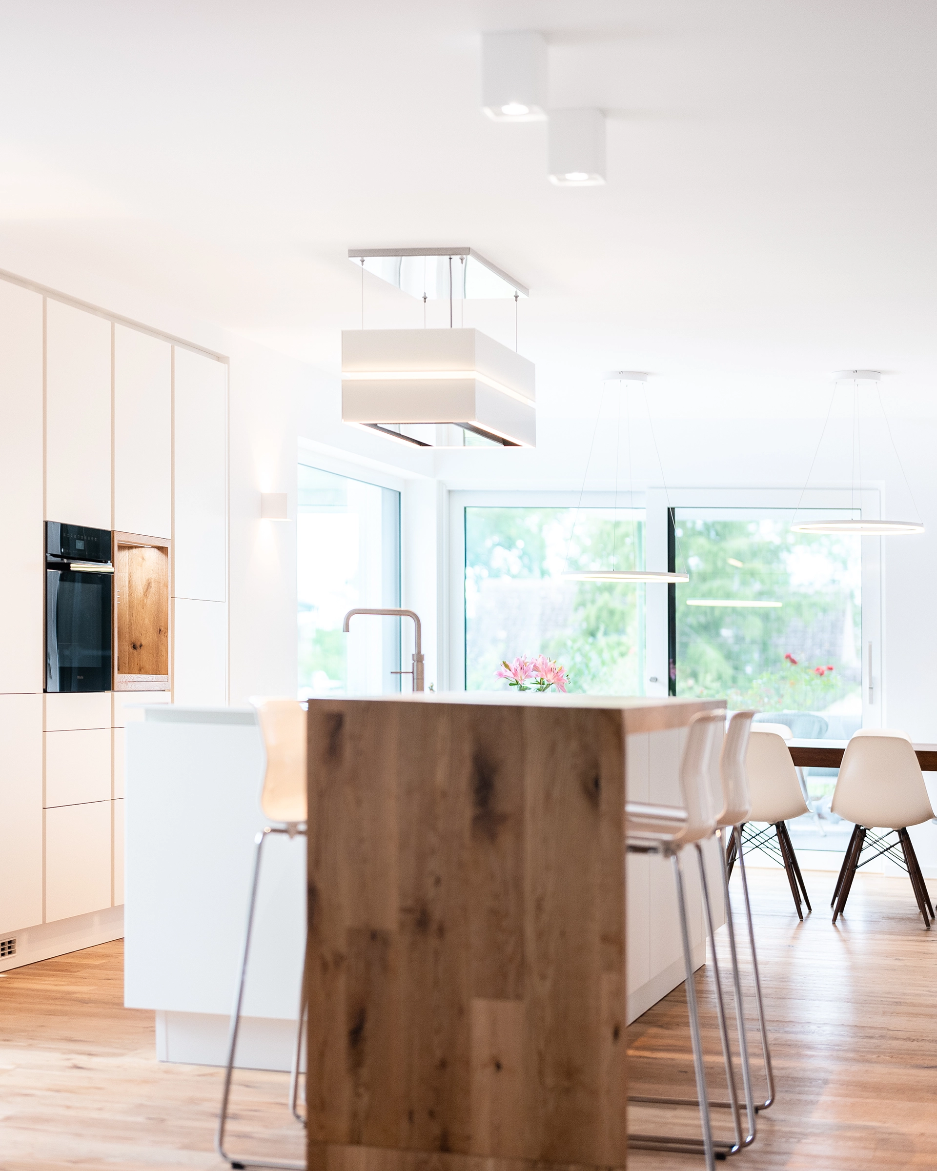 Bright modern kitchen with wooden floor, white cabinets, kitchen island with wooden end, and white bar stools.