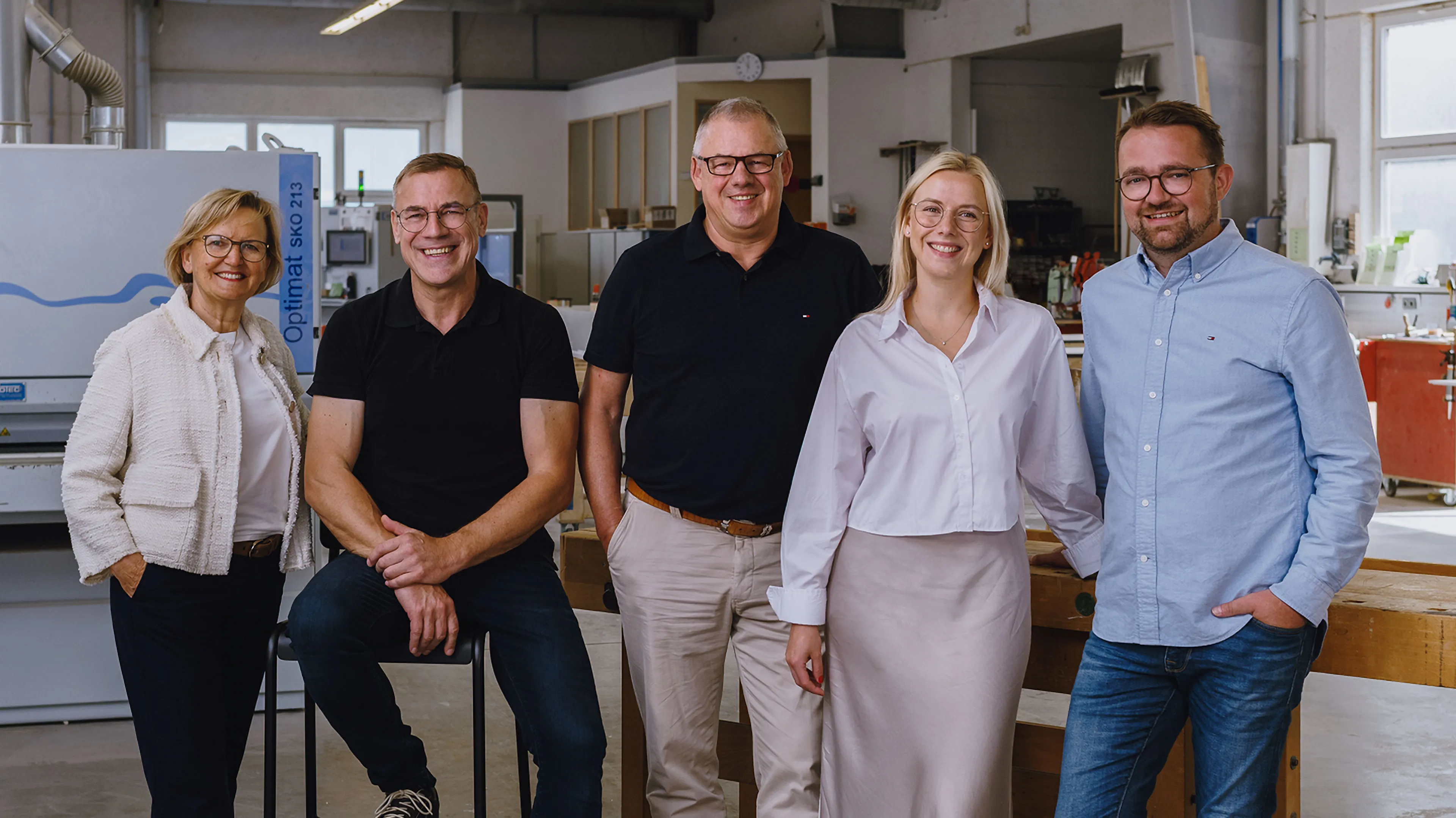 Five people smiling and posing together in an industrial workshop with machinery and wooden workbenches.
