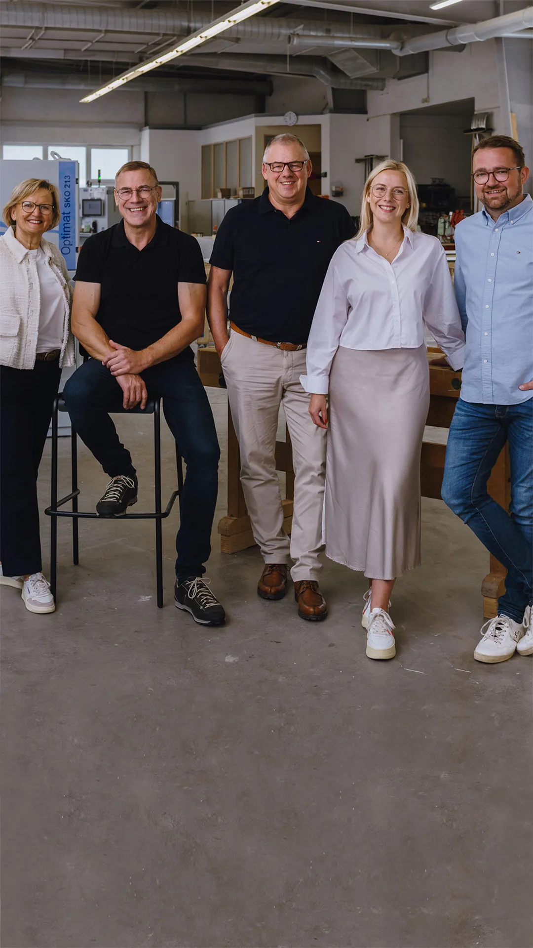 Group of five smiling people in casual business attire standing and sitting in a workshop or industrial space.