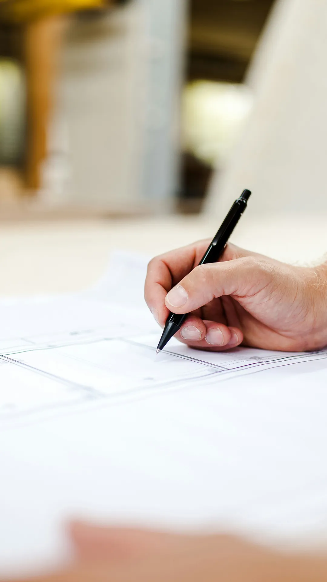 Hand holding a black pen poised over architectural blueprint or technical drawing.