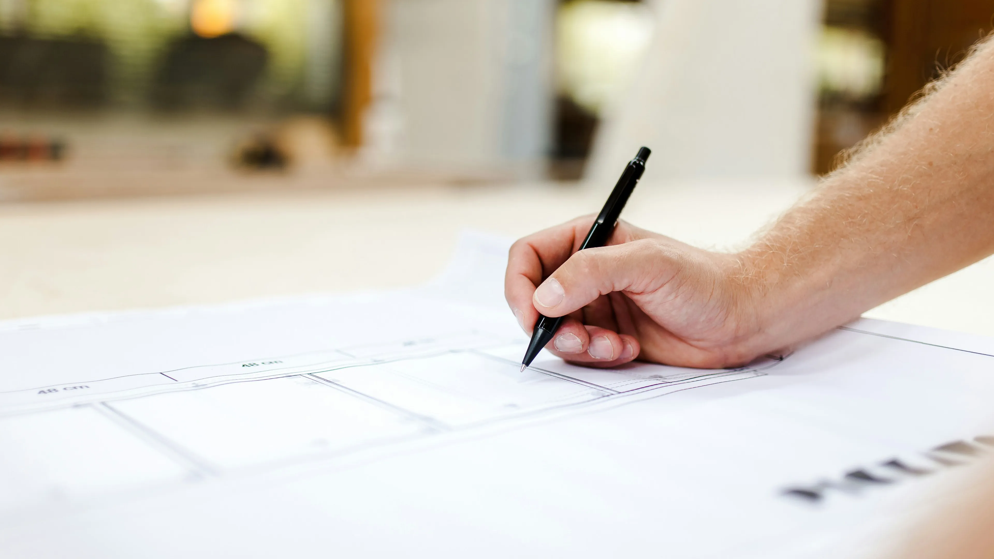 Close-up of a hand holding a black pen, drawing or writing on a large blueprint or architectural plan.