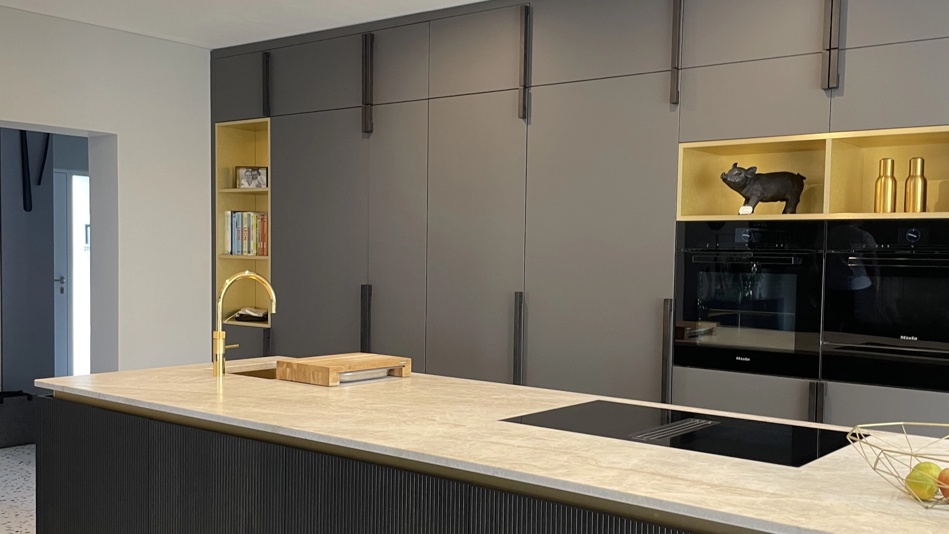 Modern kitchen with a large marble island, gold faucet, built-in black ovens, and dark grey cabinets with open shelves displaying books, a pig figurine, and gold bottles.