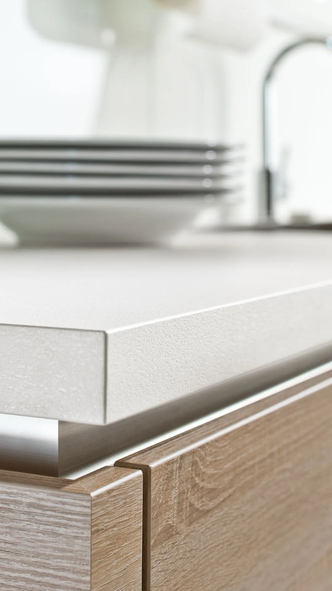 Close-up of a white textured kitchen countertop with a wooden cabinet below and stacked white plates blurred in the background.